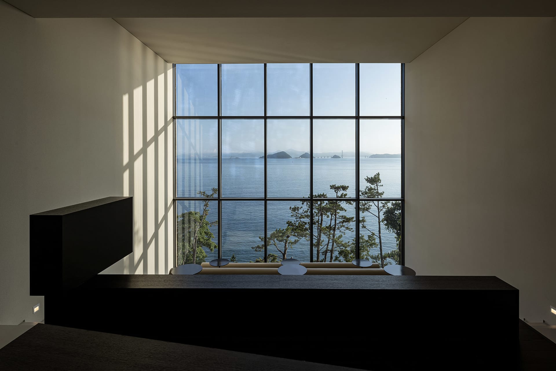 A minimalist interior features a large window overlooking the ocean, with minimal furniture and clean lines.