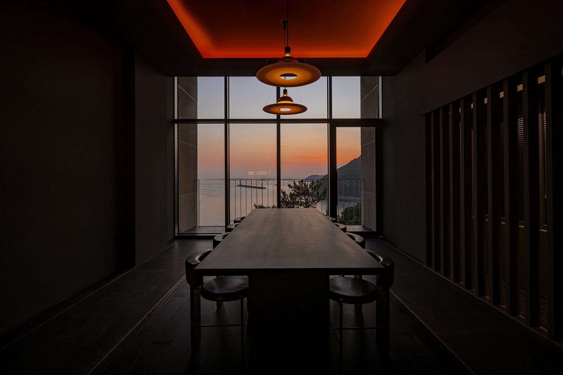 A modern dining room featuring a long wooden table and chairs, set against a large glass window overlooking a sunset over the