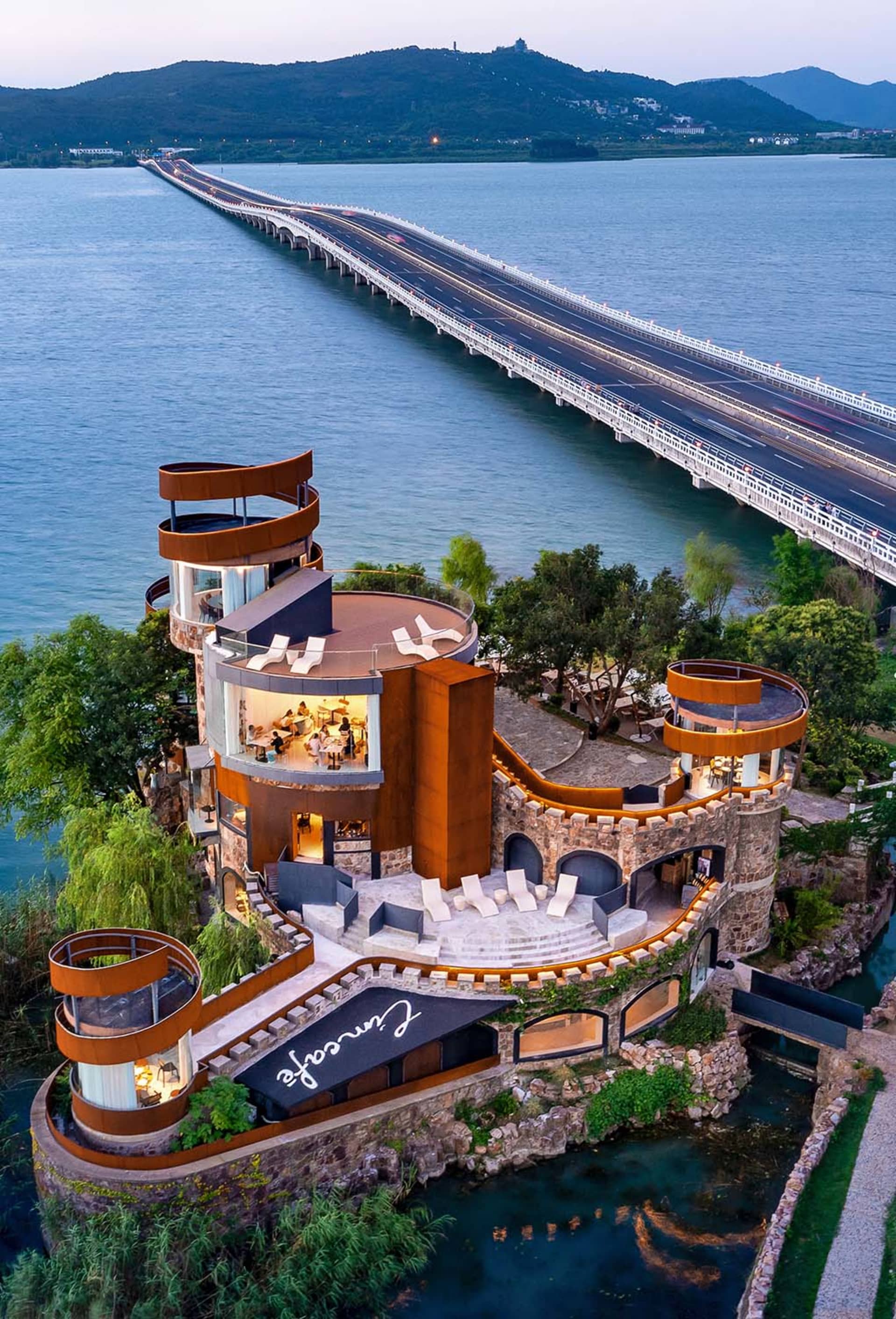 一座城堡式建築坐落於水面上,一條長橋延伸至遠方,背景為山巒。