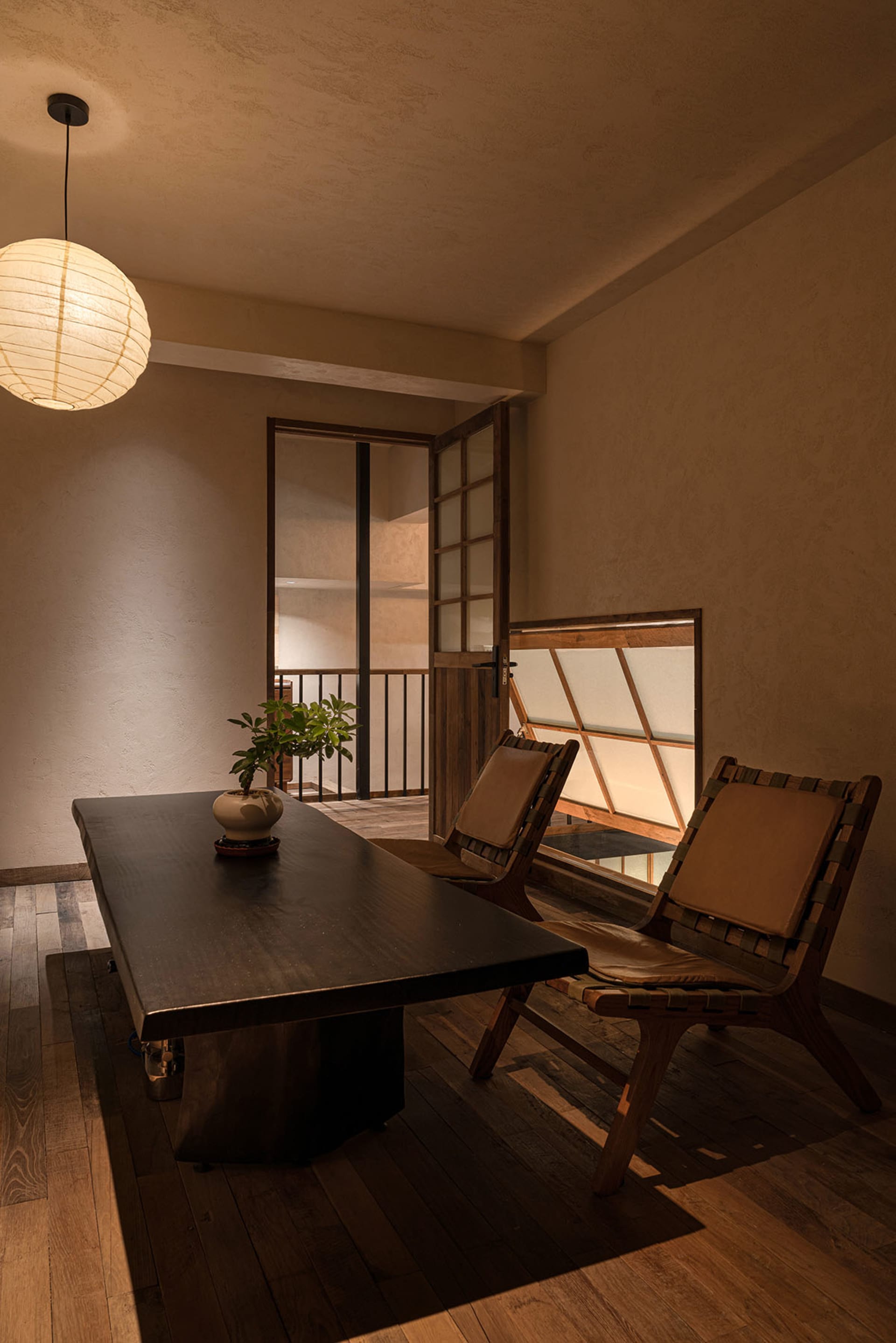 A modern minimalist office space featuring a wooden desk and chairs with a hanging lantern light.