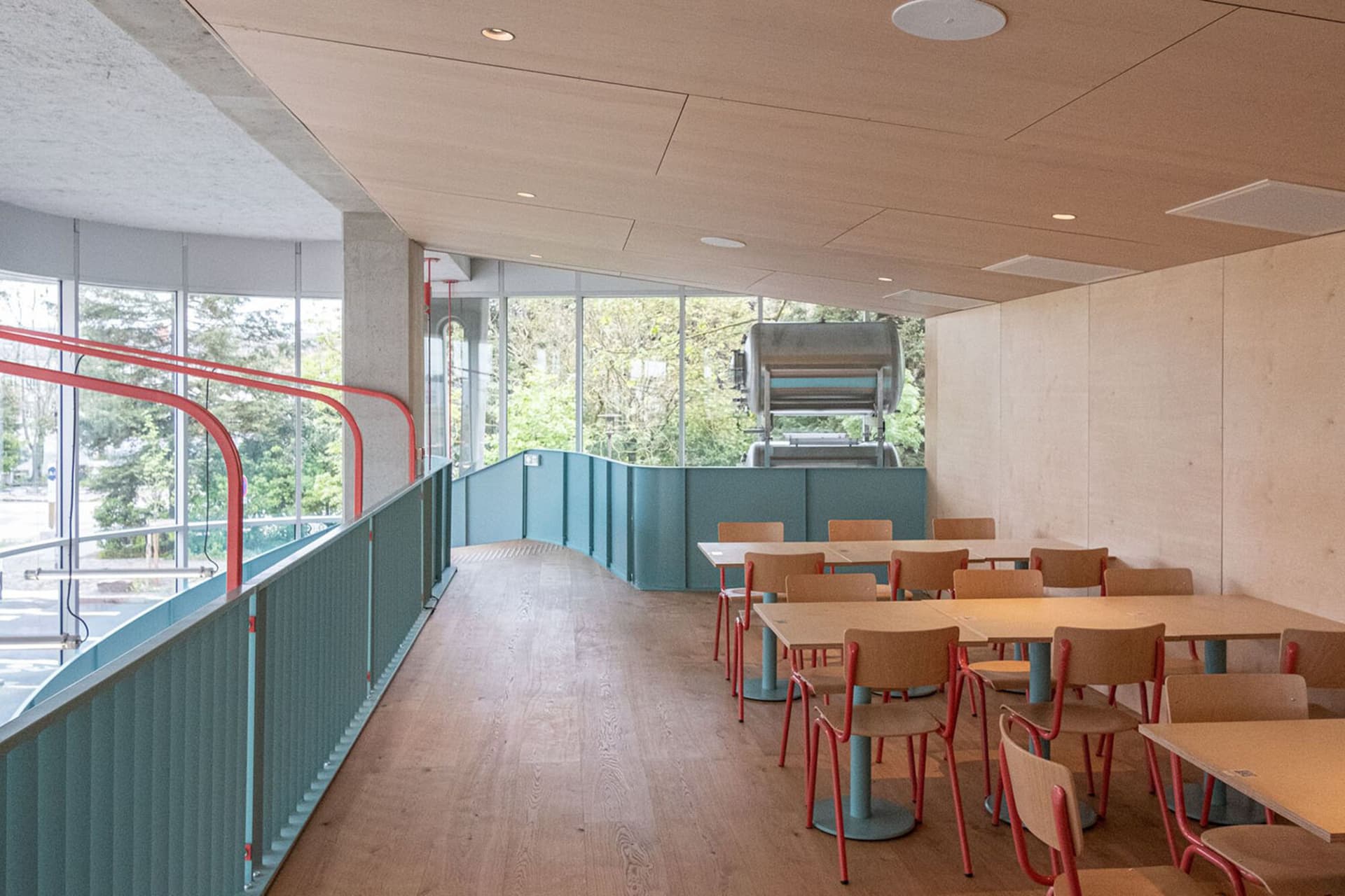 Industrial-themed restaurant interior with wooden tables and chairs, large windows and greenery outside.