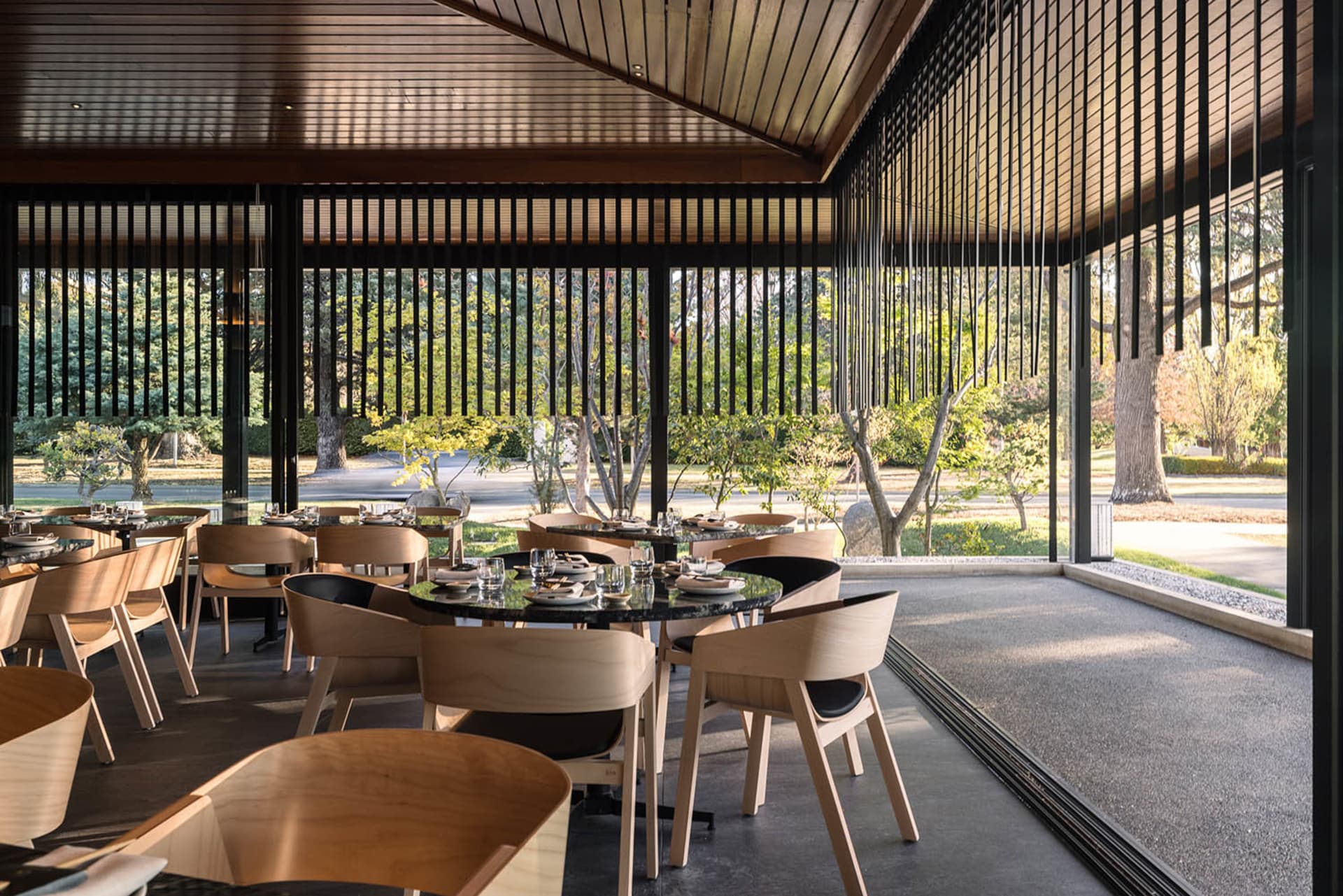 Modern restaurant interior with concrete flooring, wood furniture, and large windows overlooking greenery.