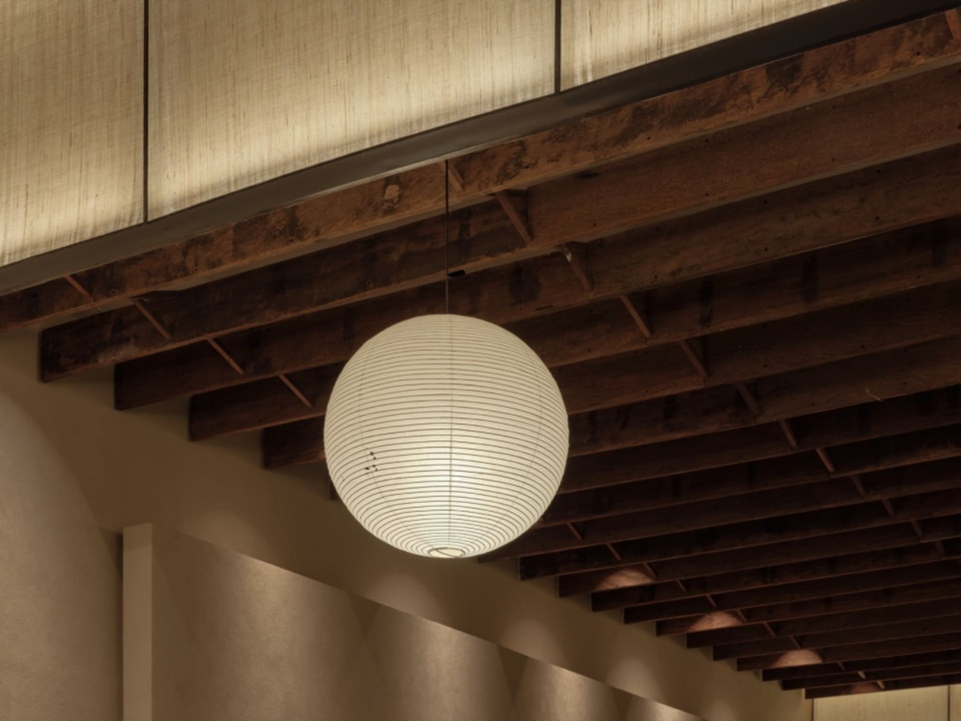A hanging spherical paper lantern illuminated from within, hanging from wooden beams in a minimalist interior setting.