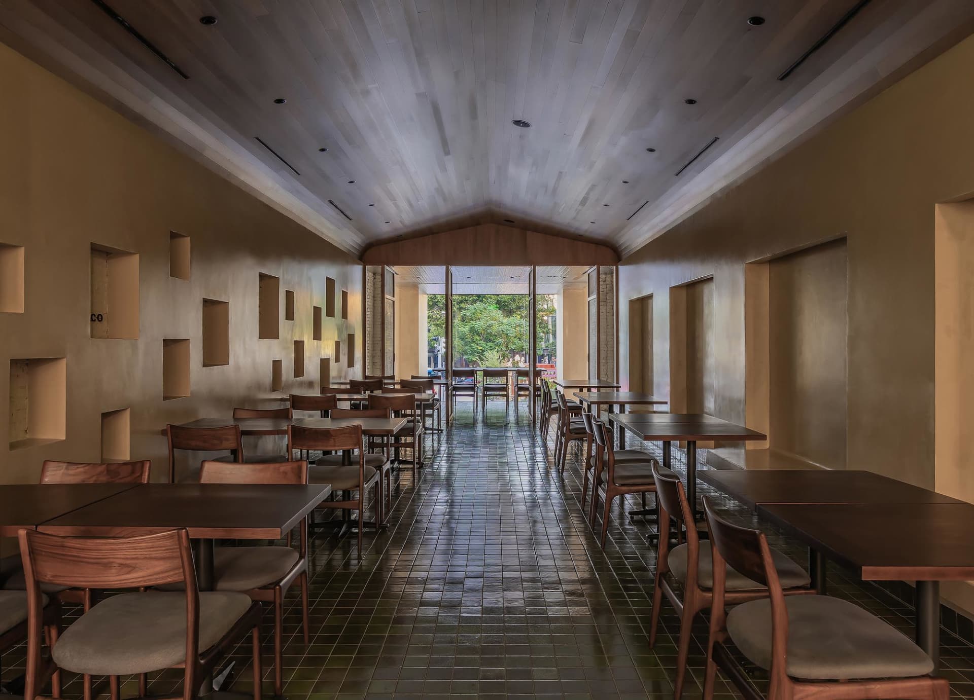 A modern restaurant interior with wooden tables and chairs, concrete walls, and a high ceiling.