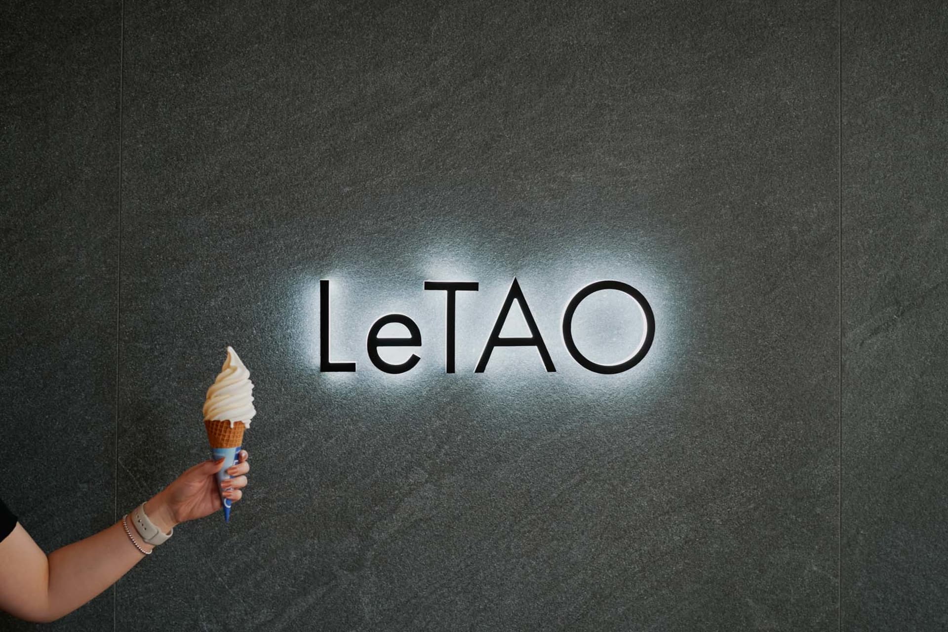 一隻手拿著冰淇淋甜筒，站在灰色大理石招牌前，背景有「LeTAO」燈光照亮。