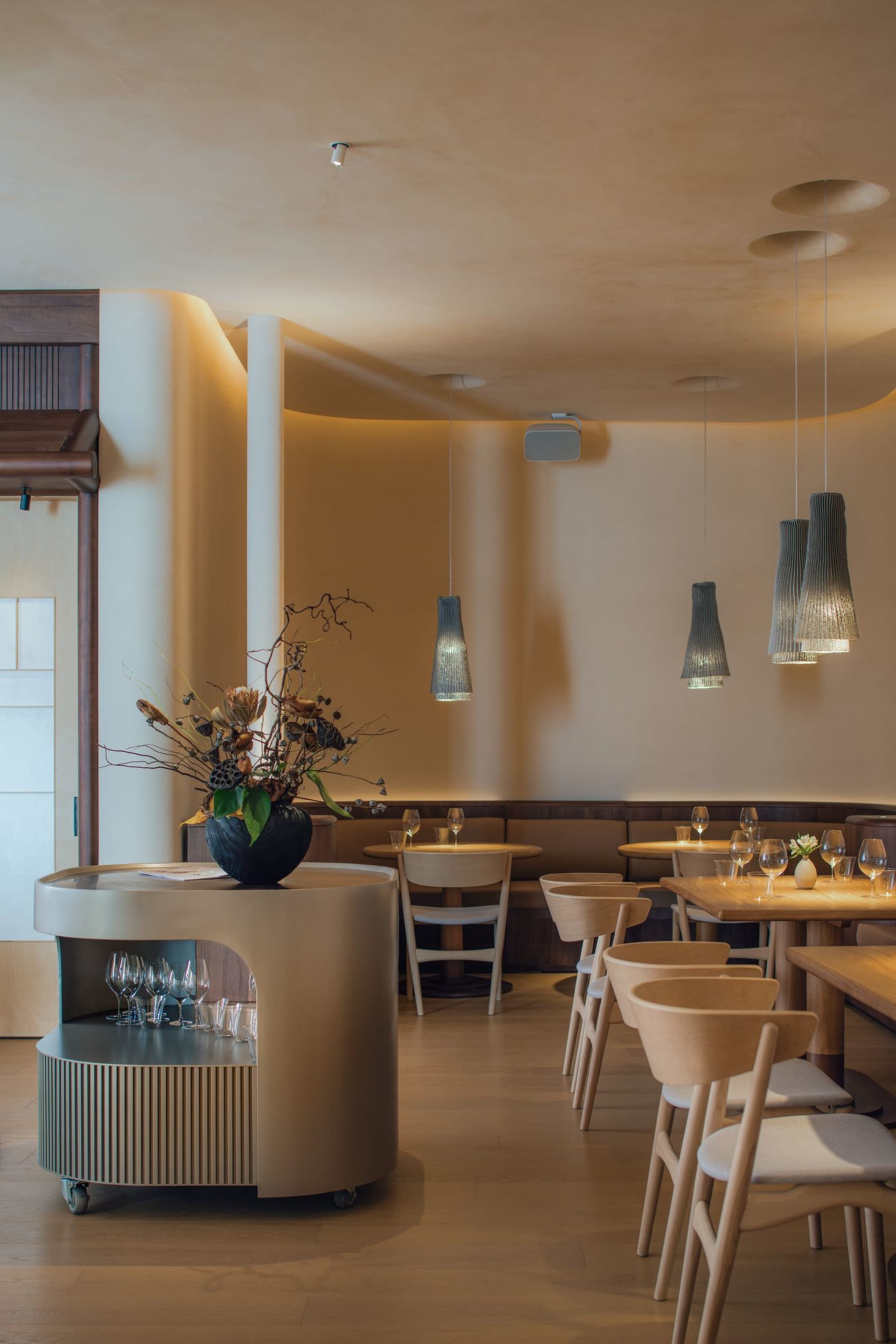 A modern restaurant interior with white walls, wooden furniture, and hanging pendant lights.