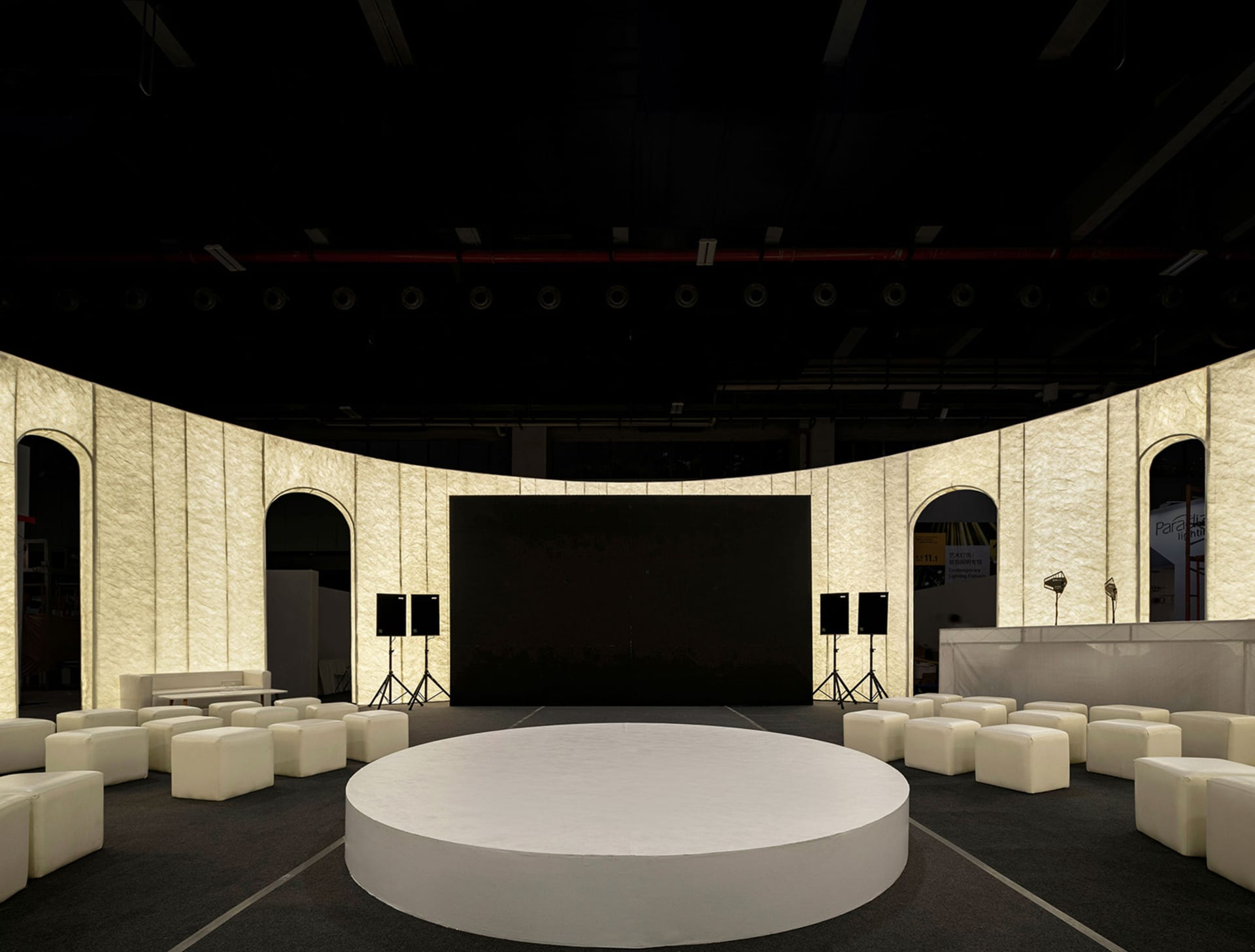 A minimalist conference room with a round, white platform at the center, surrounded by upholstered seating and concrete walls