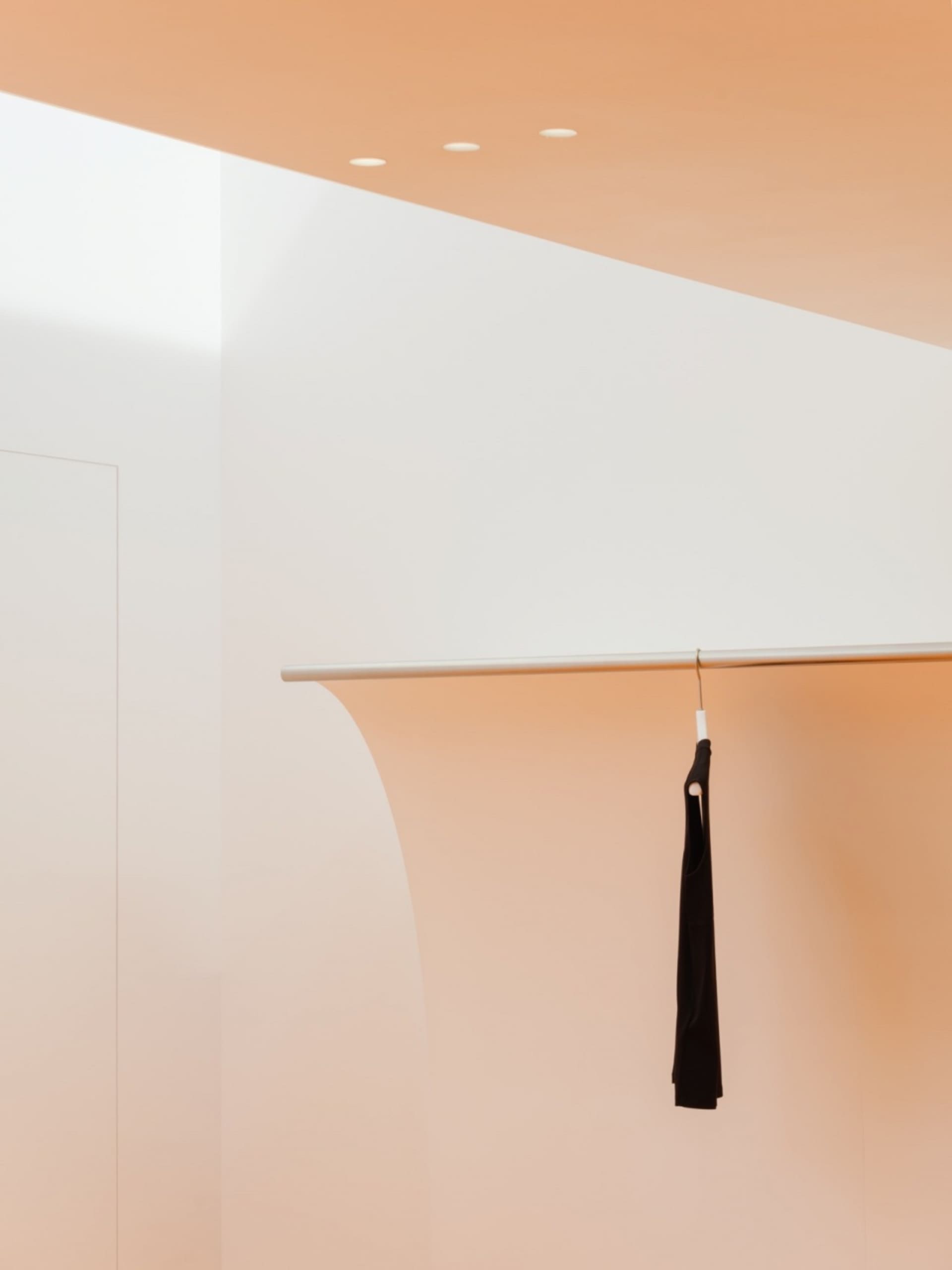 A minimalist clothing store display featuring a black garment hanging on a metallic rod against a light pink and white wall.