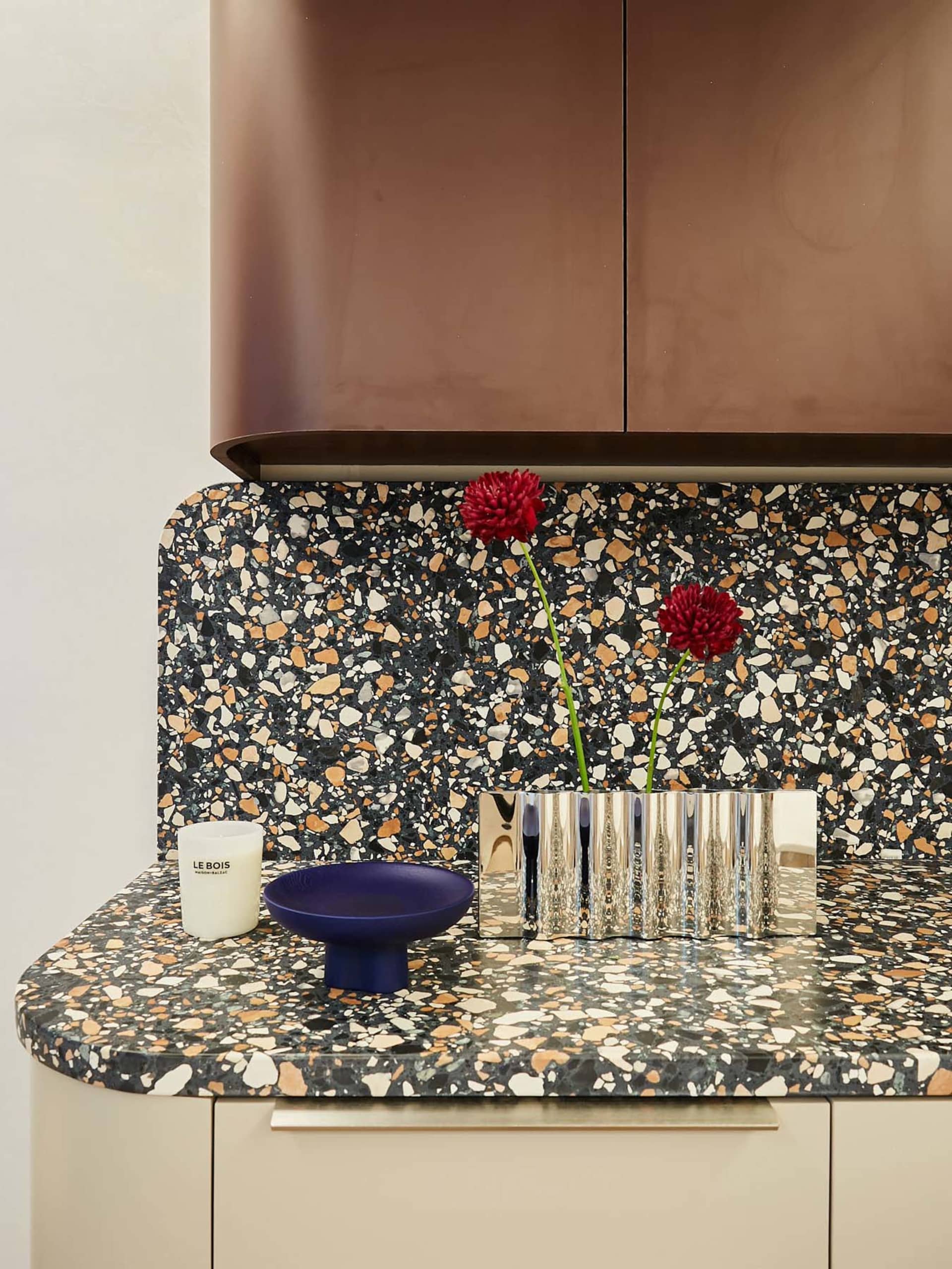 A minimalist kitchen with a colorful terrazzo countertop features a metallic vase with red flowers, a blue bowl, and a white