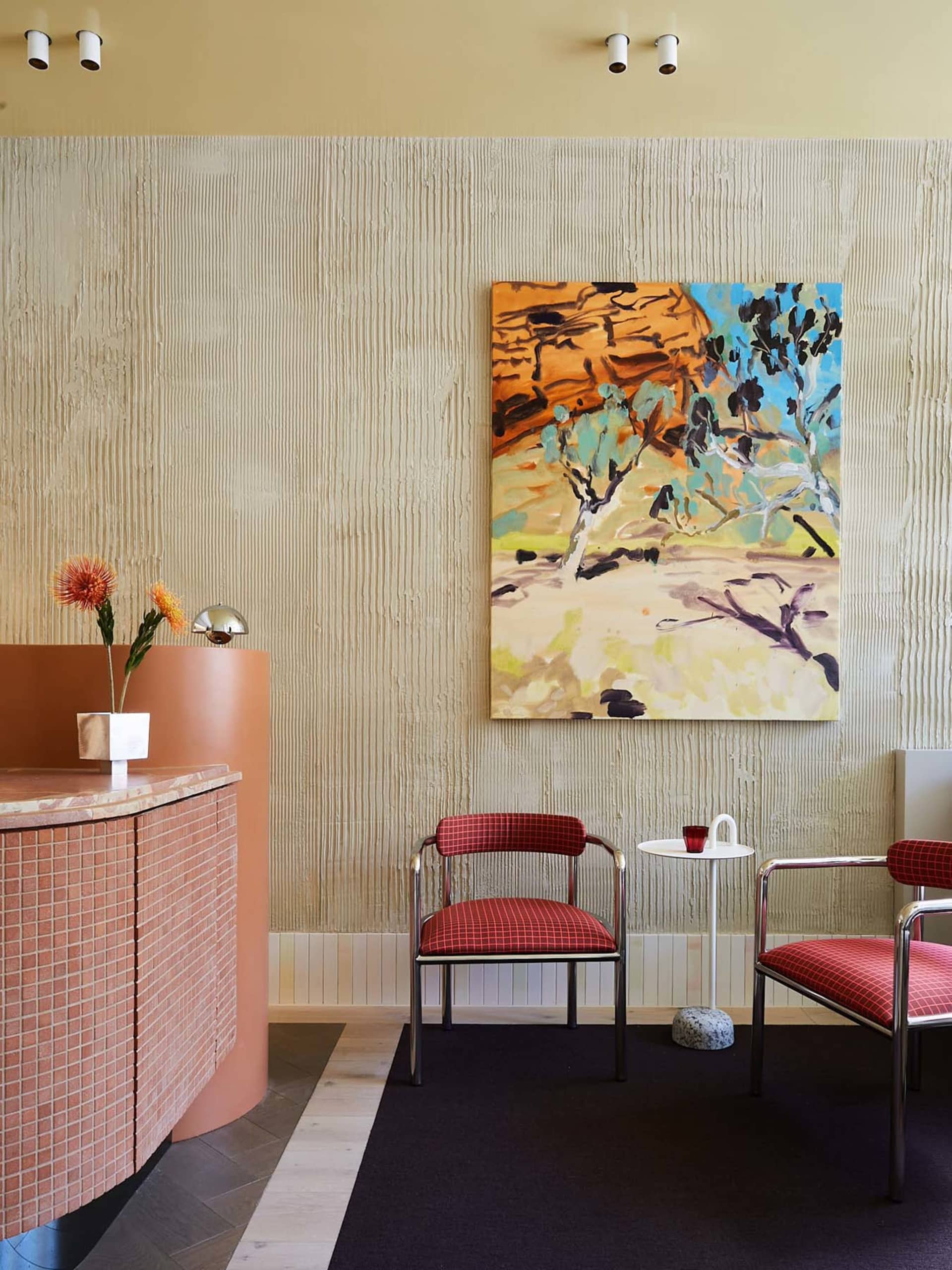 A modern restaurant lobby features a concrete reception desk with pink tiles and red cushioned chairs, alongside an abstract