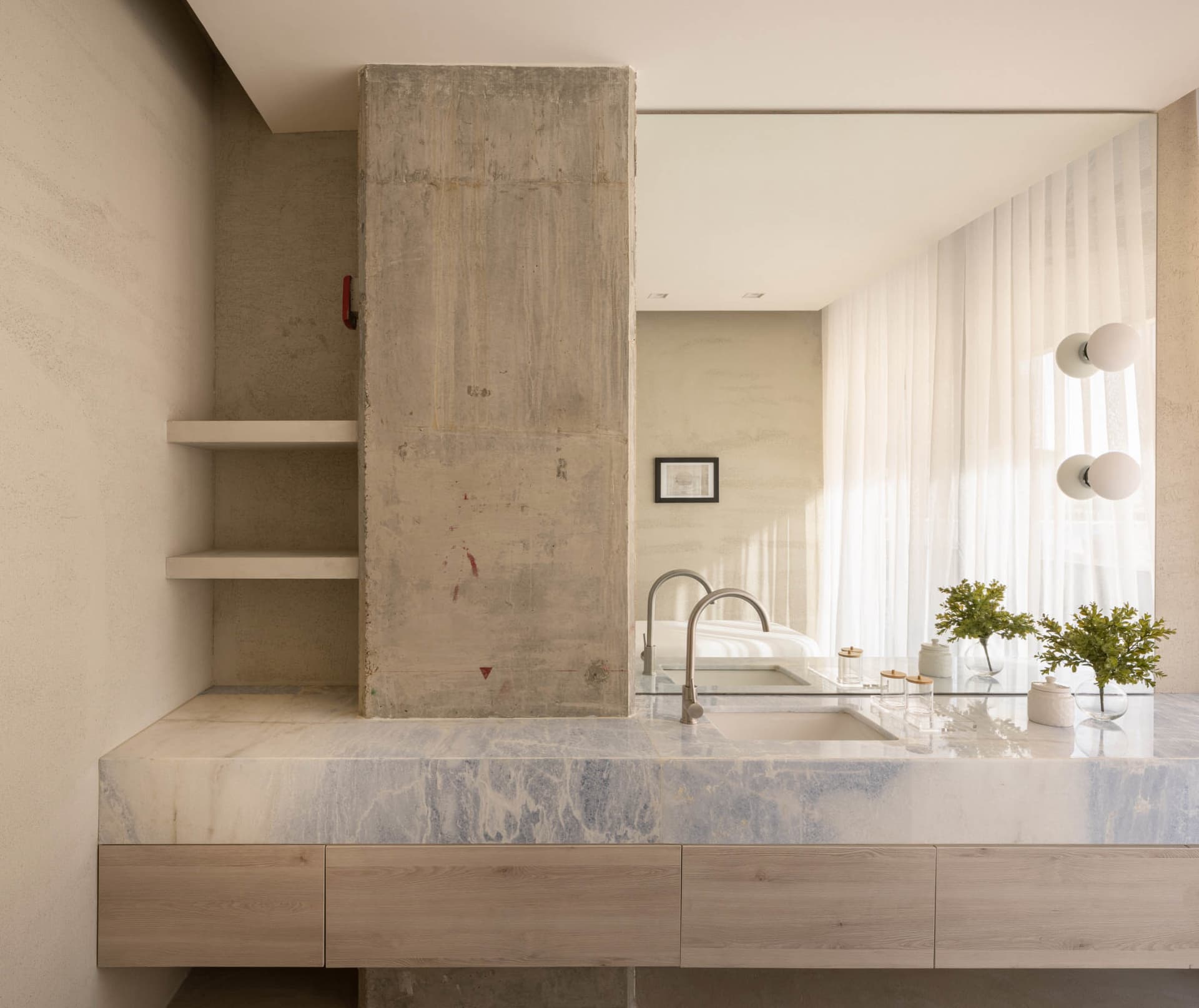 Modern minimalist bathroom featuring marble countertop, concrete partition and wooden drawers, with a view of a bedroom.