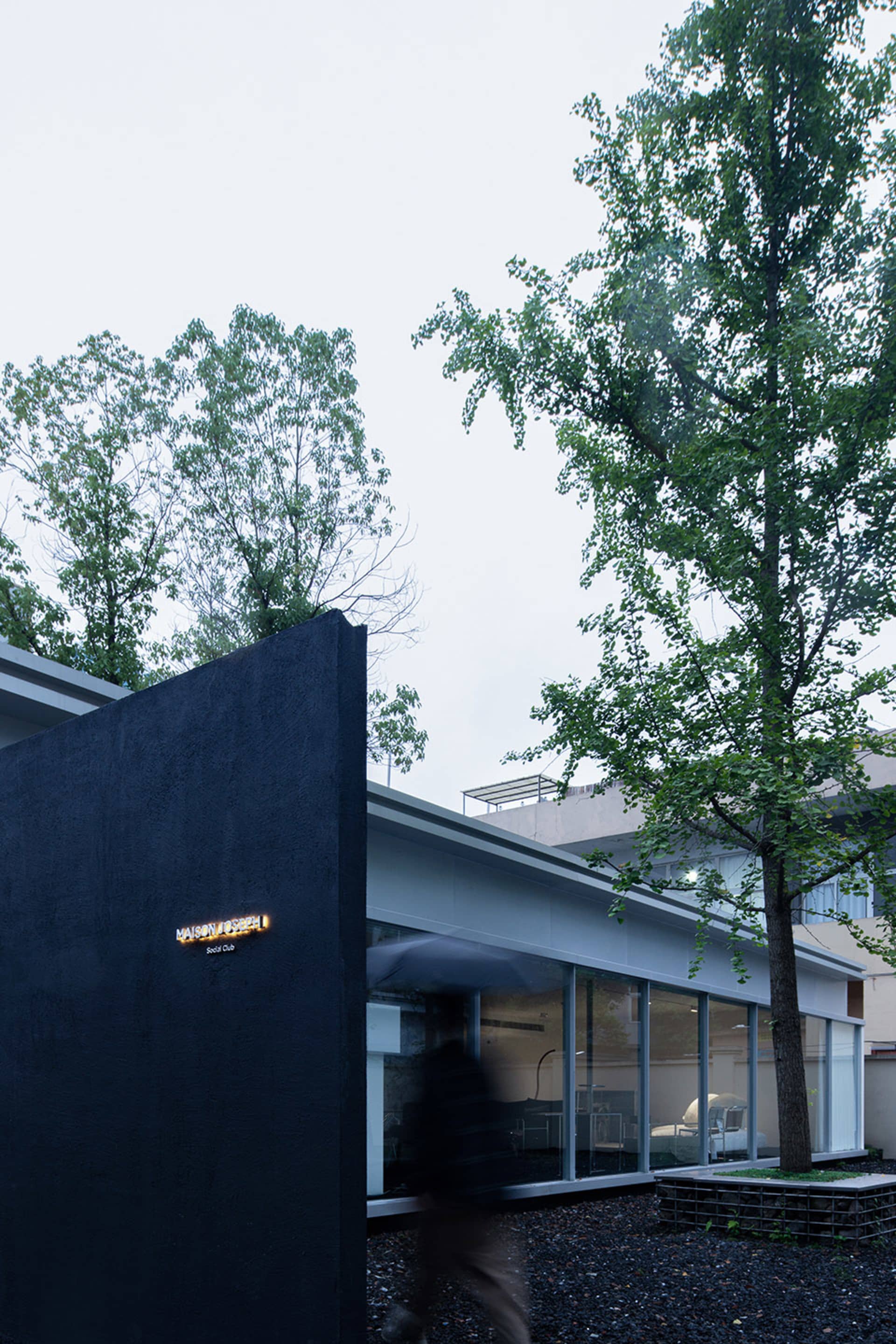 A modern social club with a black wall, glass windows, and steel accents, surrounded by green trees and gravel.