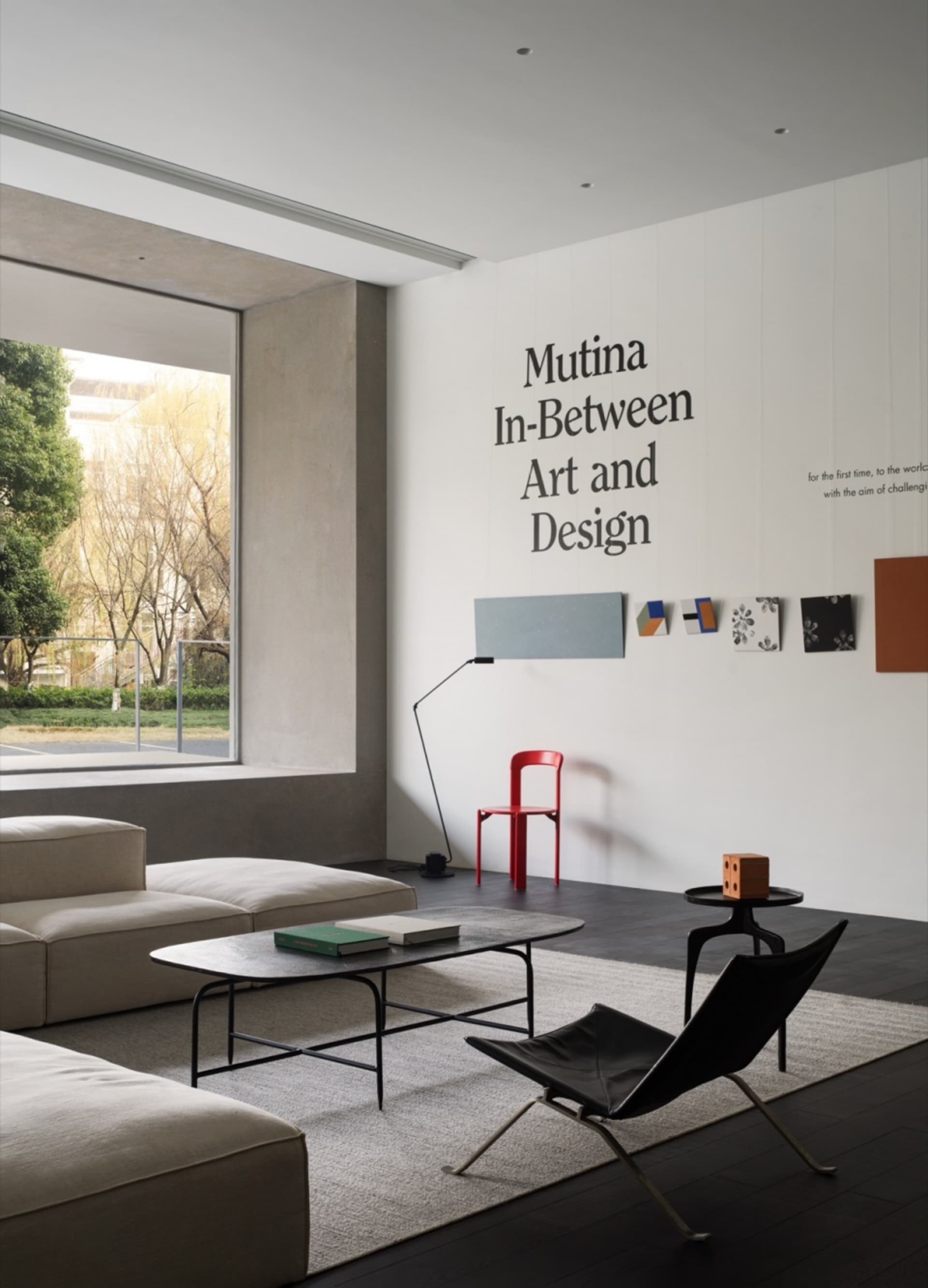 Modern minimalist living room with concrete, leather, and metal furniture, large window, and wall text reading 'Mutina In-Bet