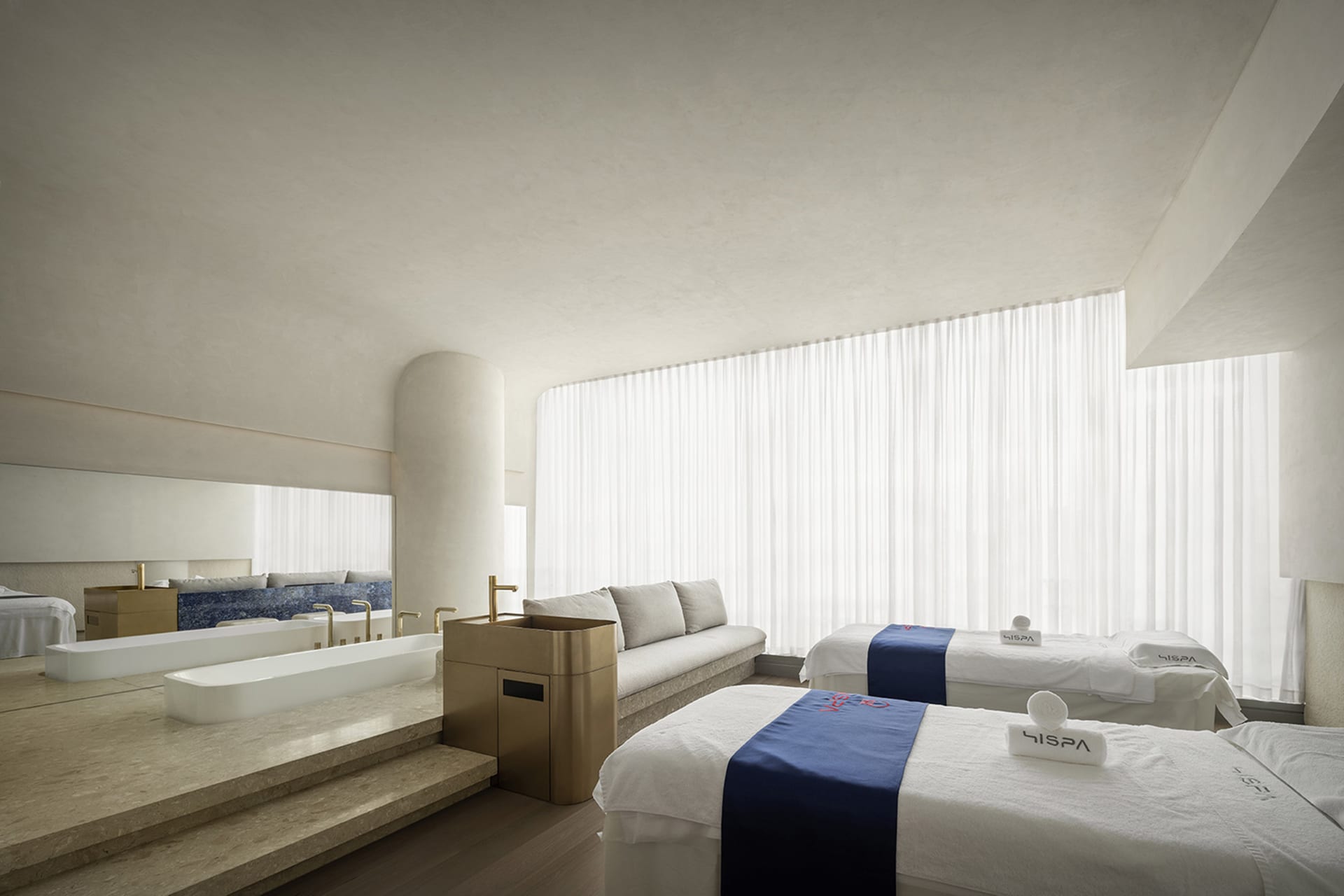 A serene spa room with white cloth textiles on stone flooring, glass walls, and a large window.