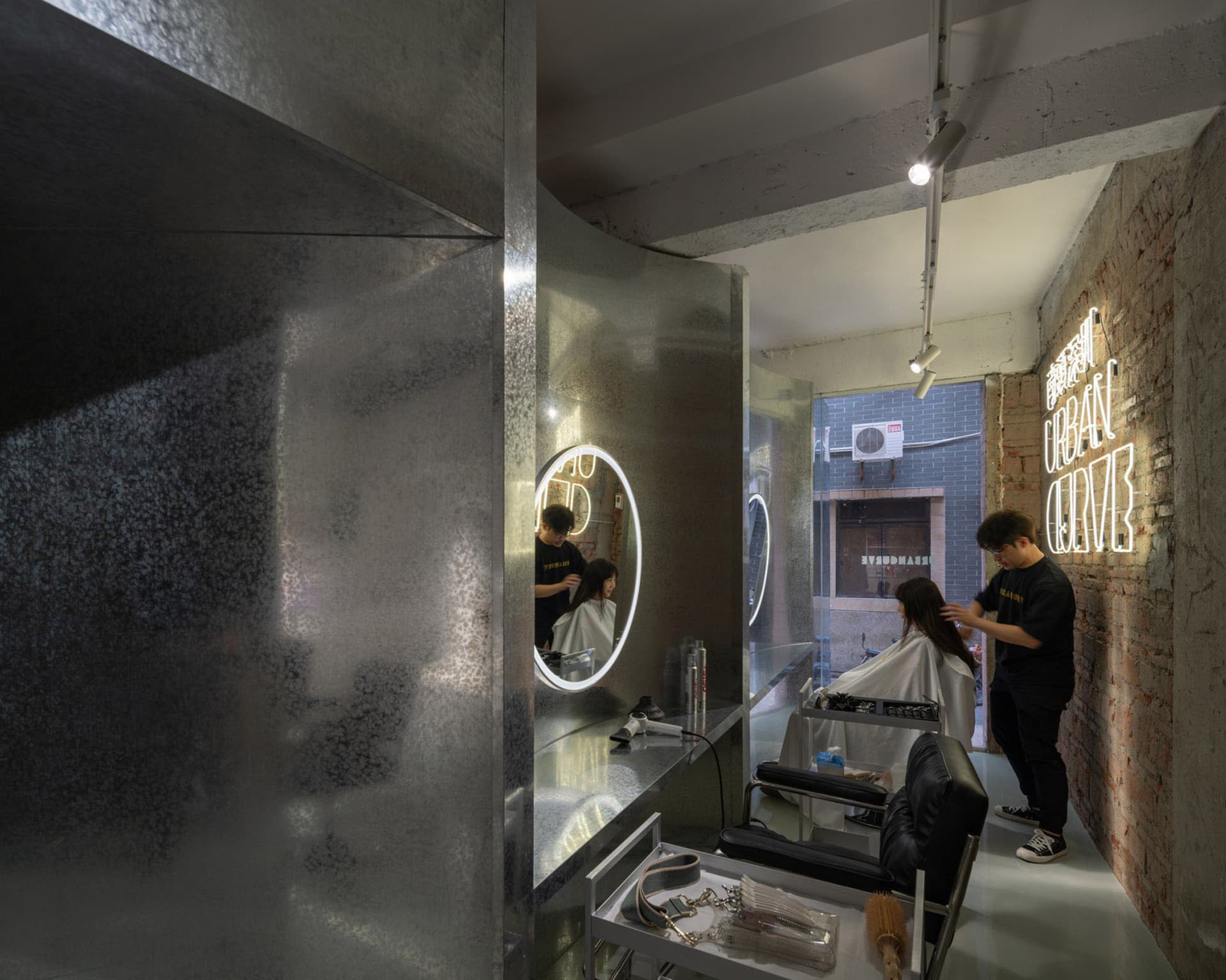現代美髮沙龍,擁有金屬牆面、磚塊點綴與混凝土地板。造型師正在為客戶整理頭髮。