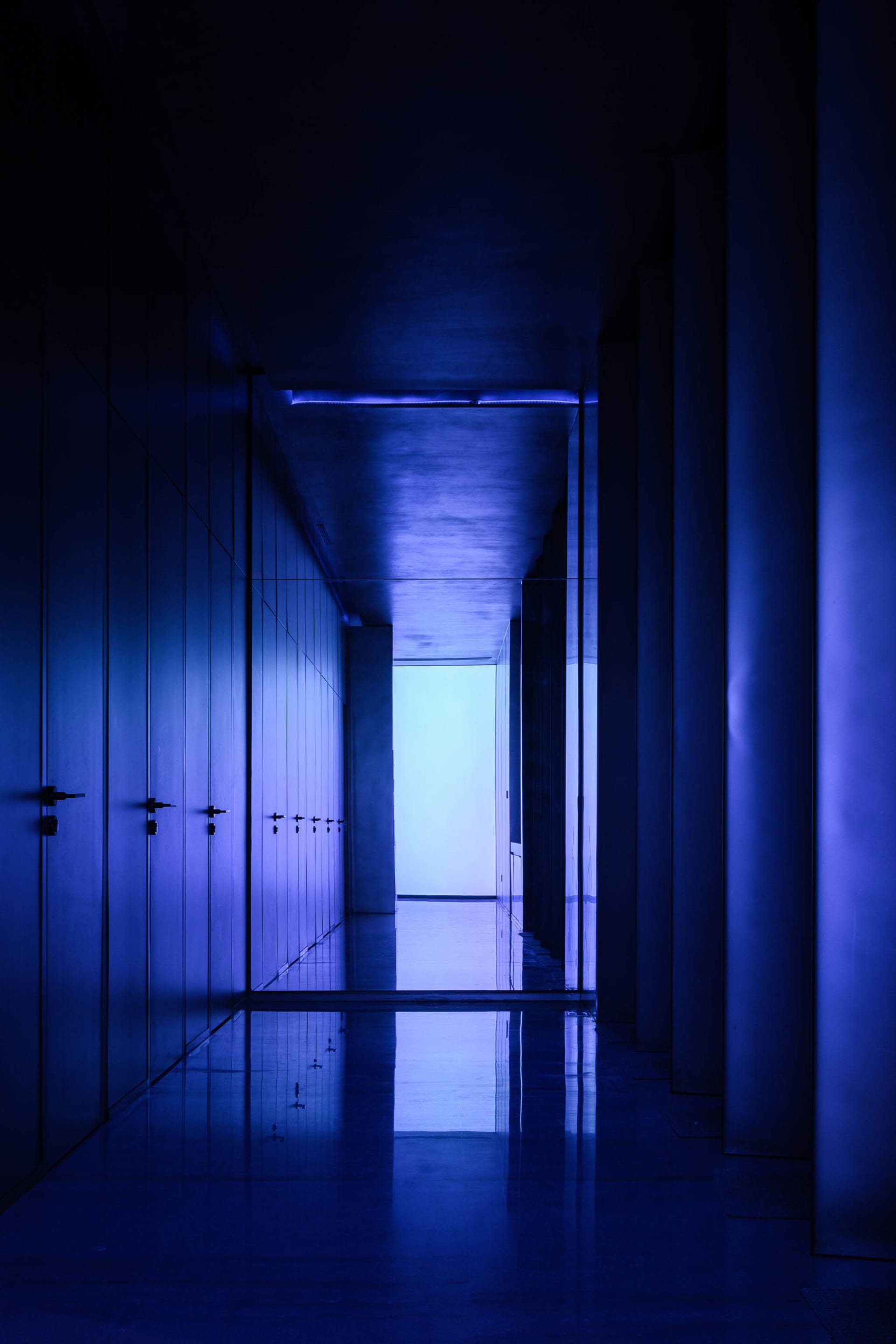 A long, narrow corridor with walls illuminated in a single, deep blue hue, extending towards a bright endpoint.