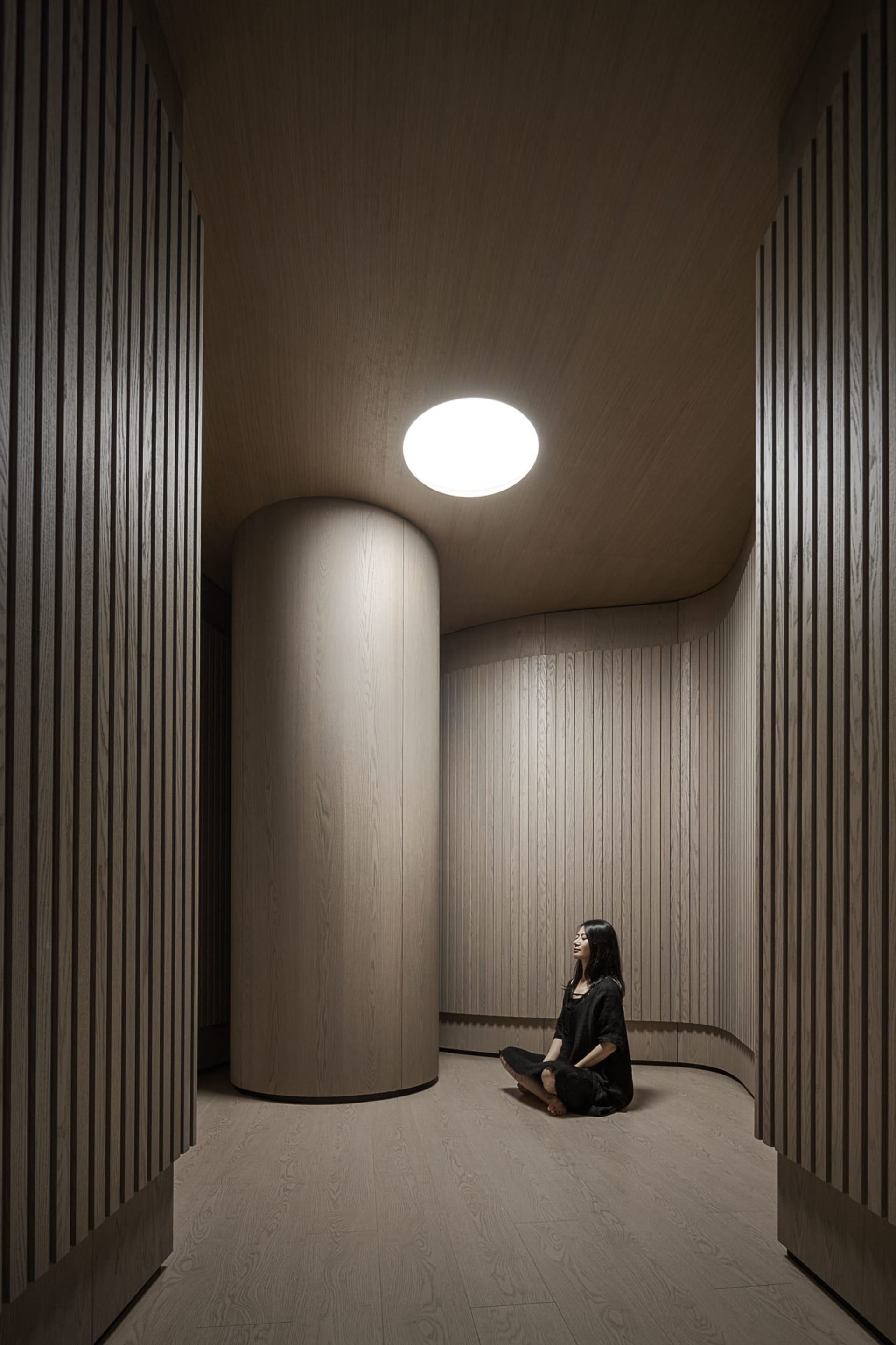 A minimalist living room with wooden walls and floors, featuring a woman sitting in a serene pose.