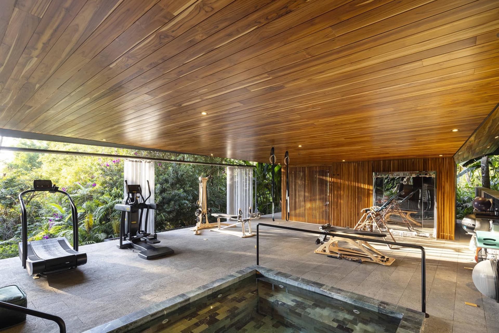A modern home gym with wooden flooring and ceiling, featuring exercise equipment and a pool area.