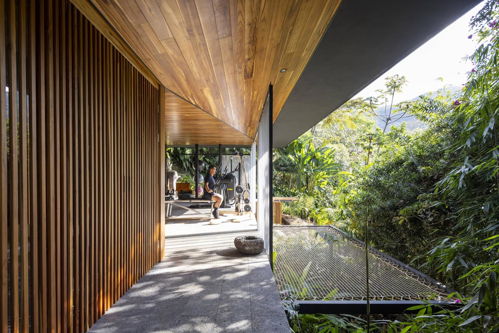 An outdoor gym with wooden flooring and walls, featuring exercise equipment and surrounded by lush greenery.