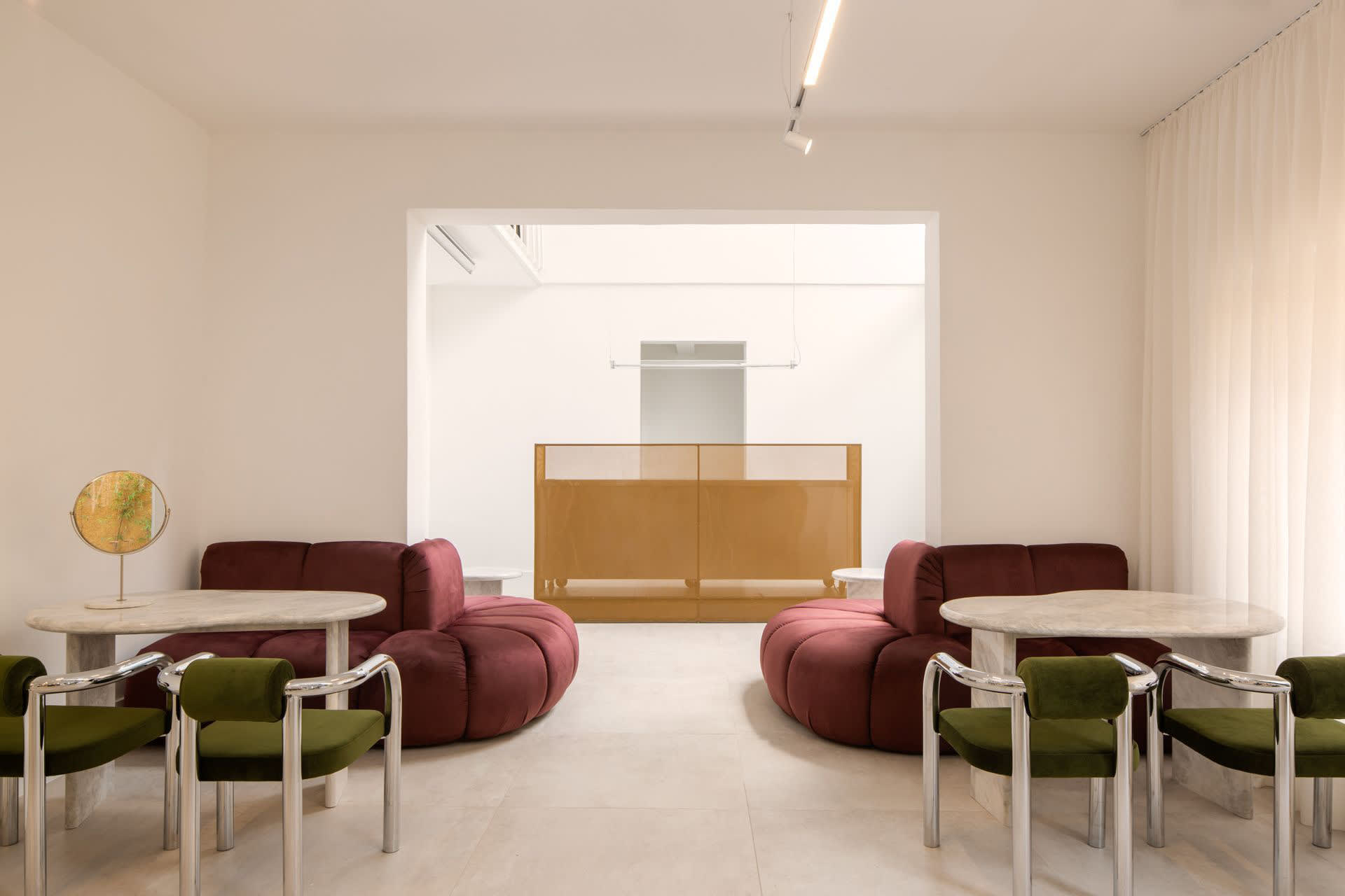 A minimalist living room featuring two round, red sofas, green chairs, marble tables, and a gold-toned divider.
