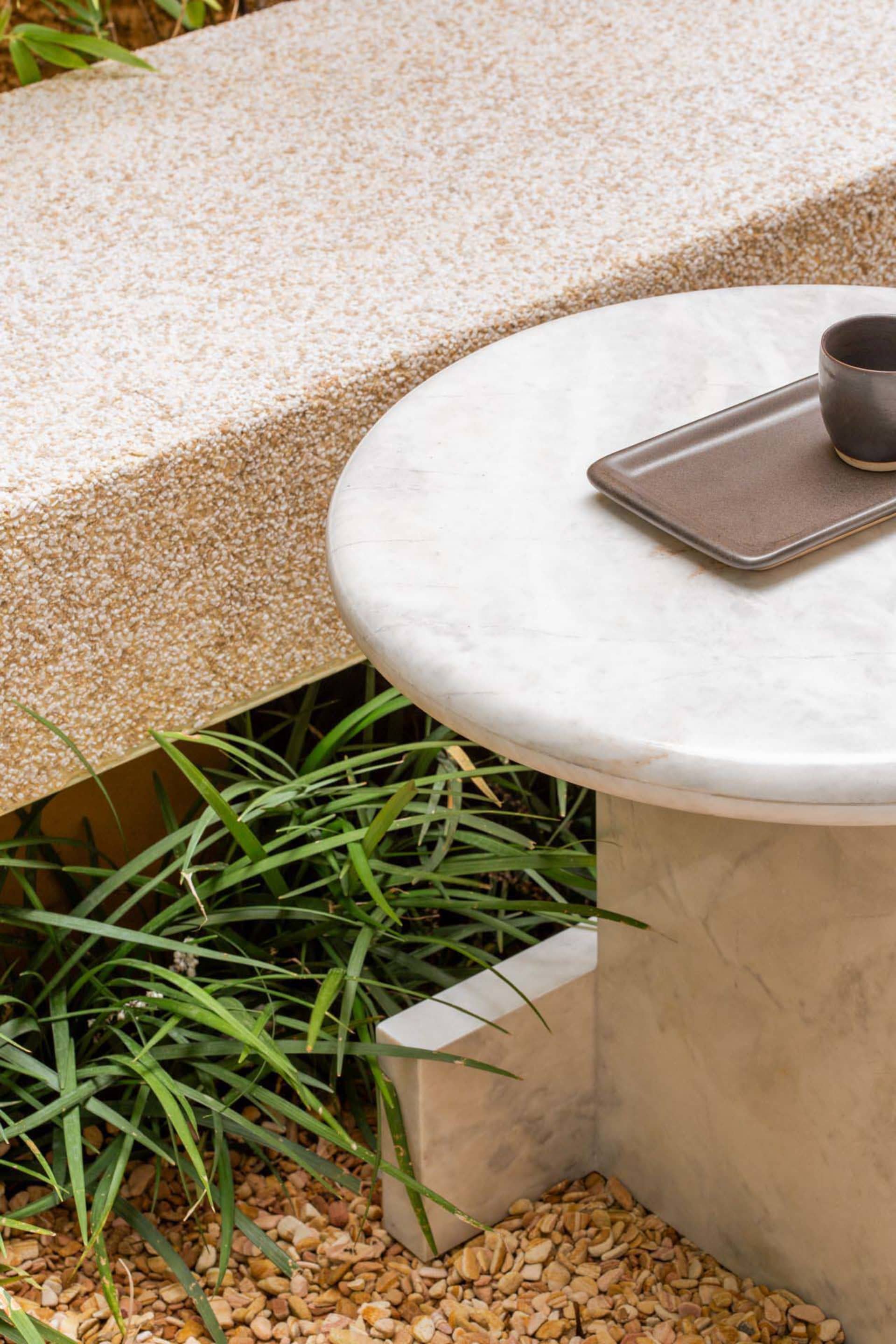 A round marble table with a tray and cup on a natural stone patio surrounded by greenery.