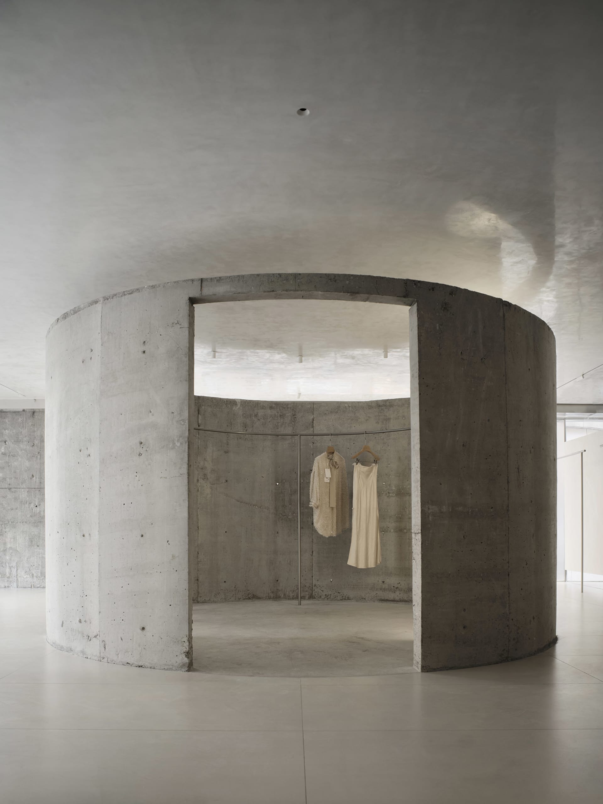A minimalist fashion retail store with concrete walls and a concrete floor, featuring beige clothing displayed on hangers.