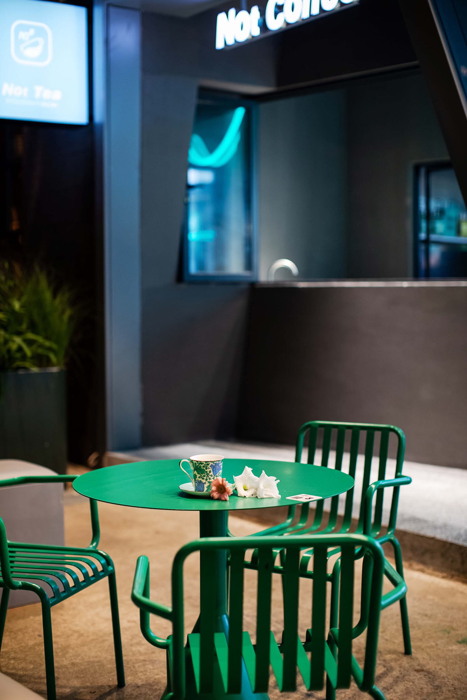 A minimalistic café's espresso bar setup featuring green metal furniture, a floral teacup, and white flowers.