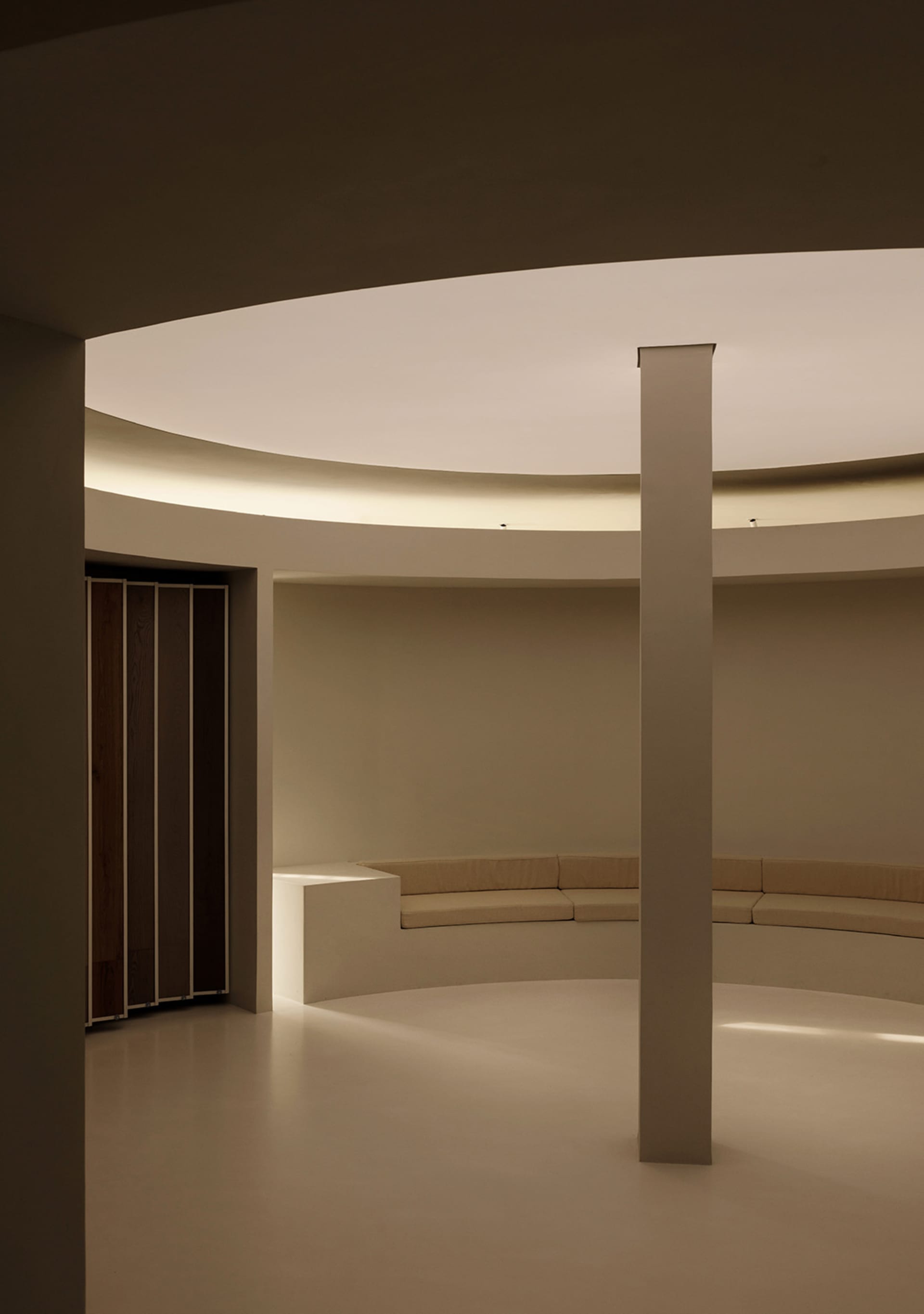 A minimalist interior space with a circular seating area and a central pillar, made of concrete.