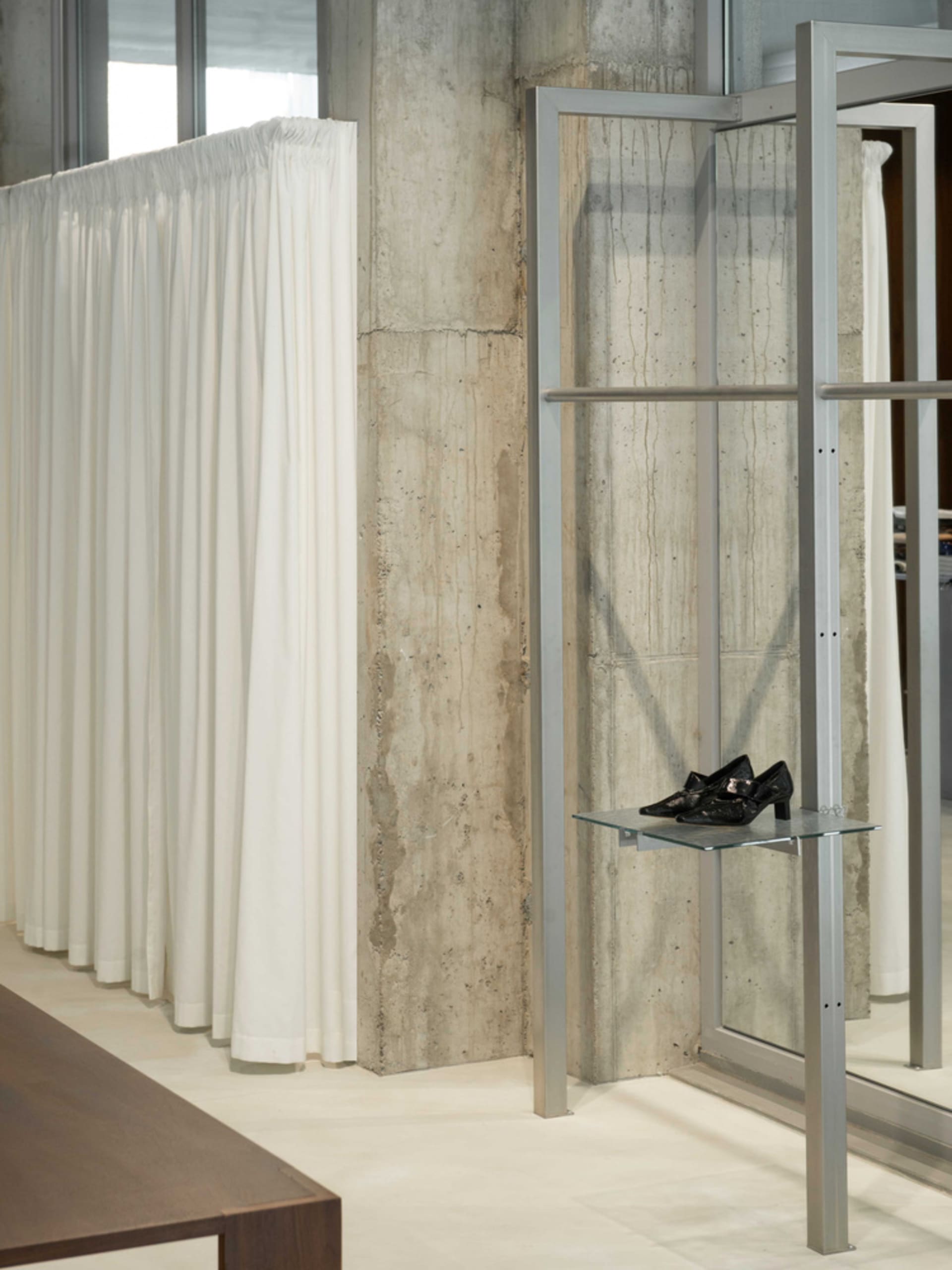 A modern fashion shop featuring a concrete wall, white curtains, and black shoes arranged on an aluminum shelf.