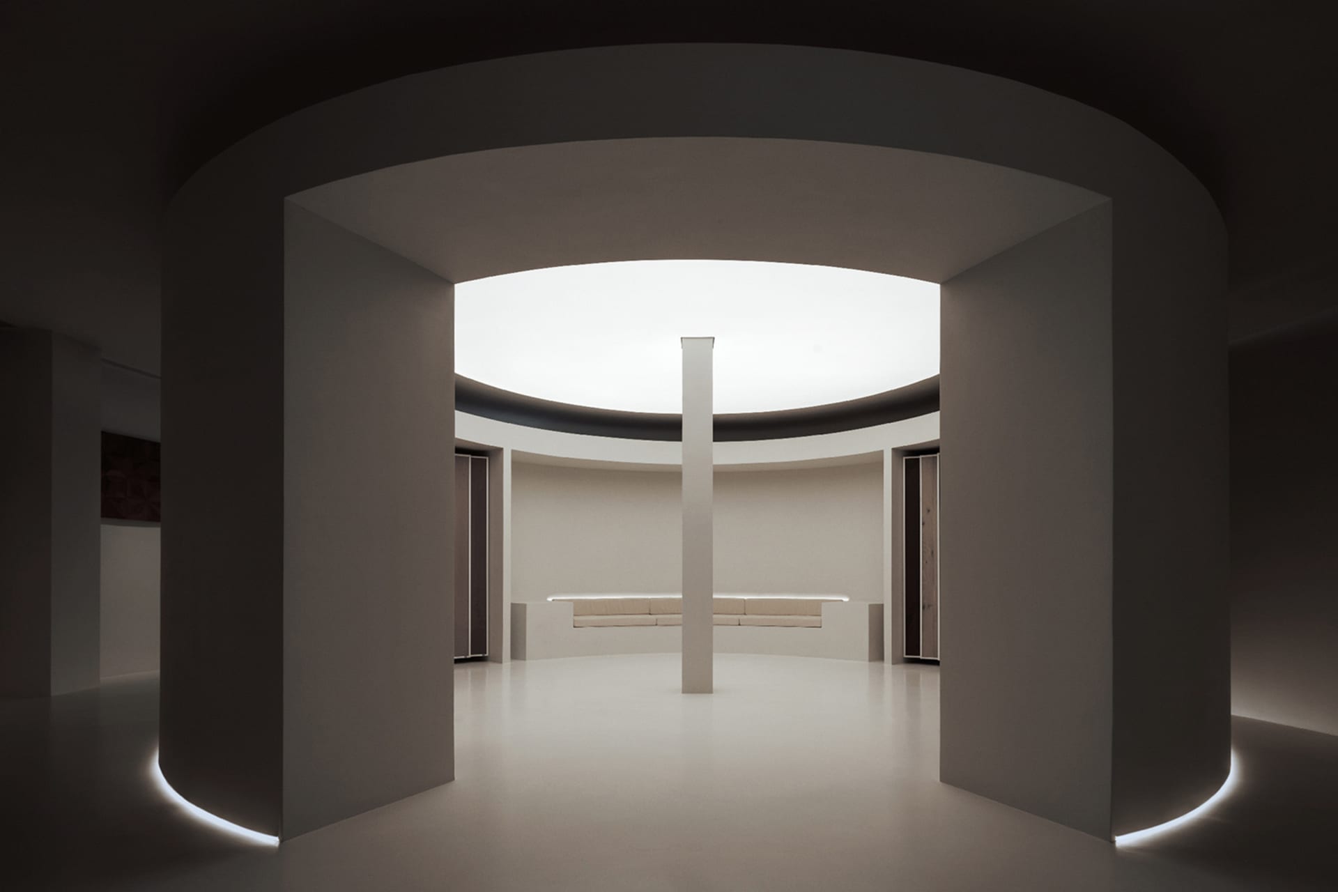 Modern minimalist living room with white and concrete surfaces, highlighted by a central column and circular light fixture.