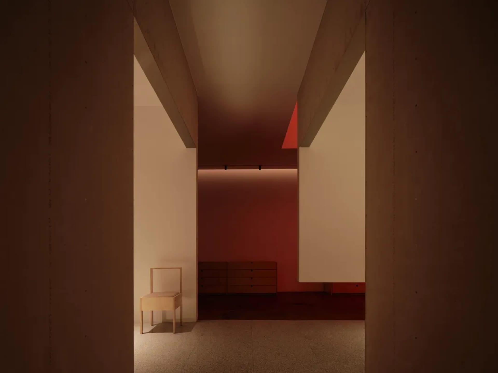 A minimalist bedroom with concrete walls and floor, featuring a simple wooden chair and a low cabinet.