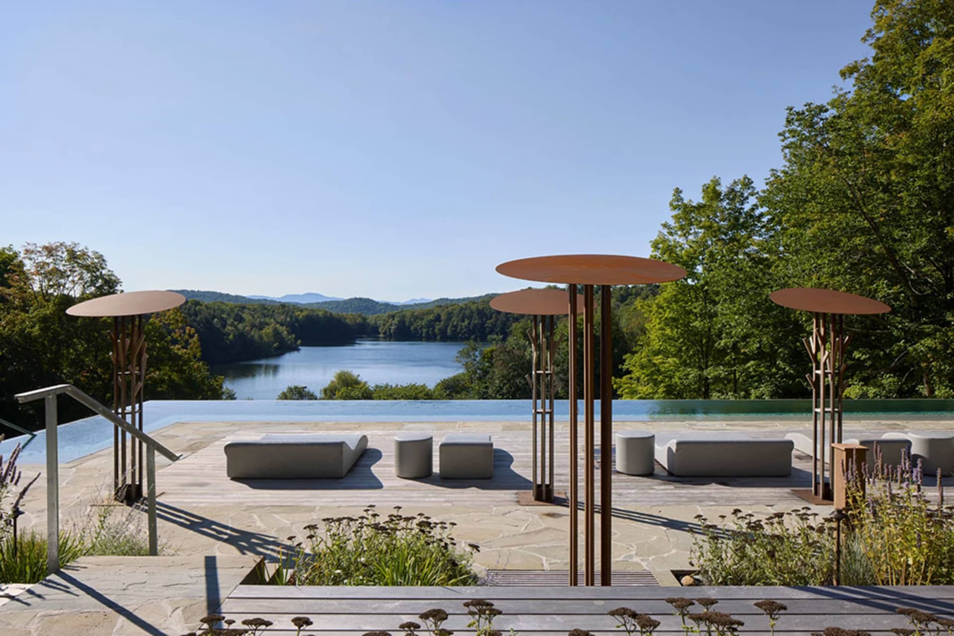 A modern outdoor living space featuring a wooden deck, infinity pool, and scenic lake view surrounded by lush greenery.
