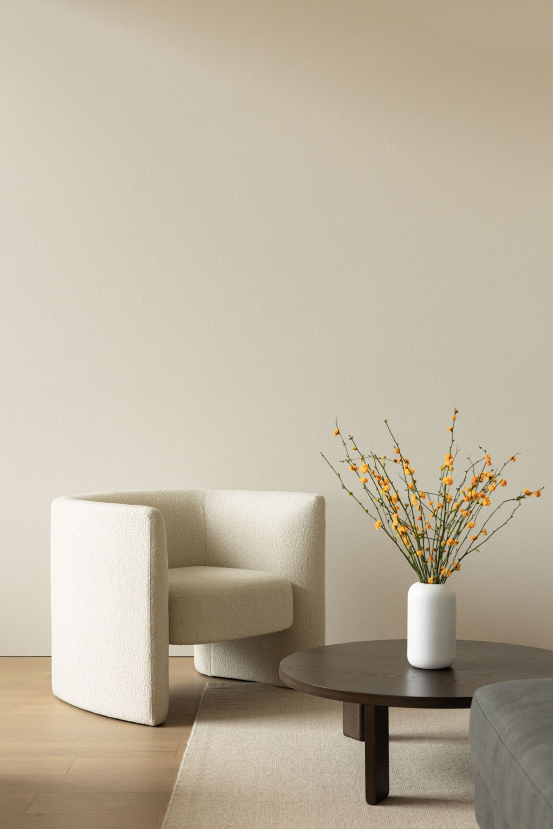 A minimalist living room with a white textured fabric armchair, round wooden coffee table, and a vase with yellow flowers.