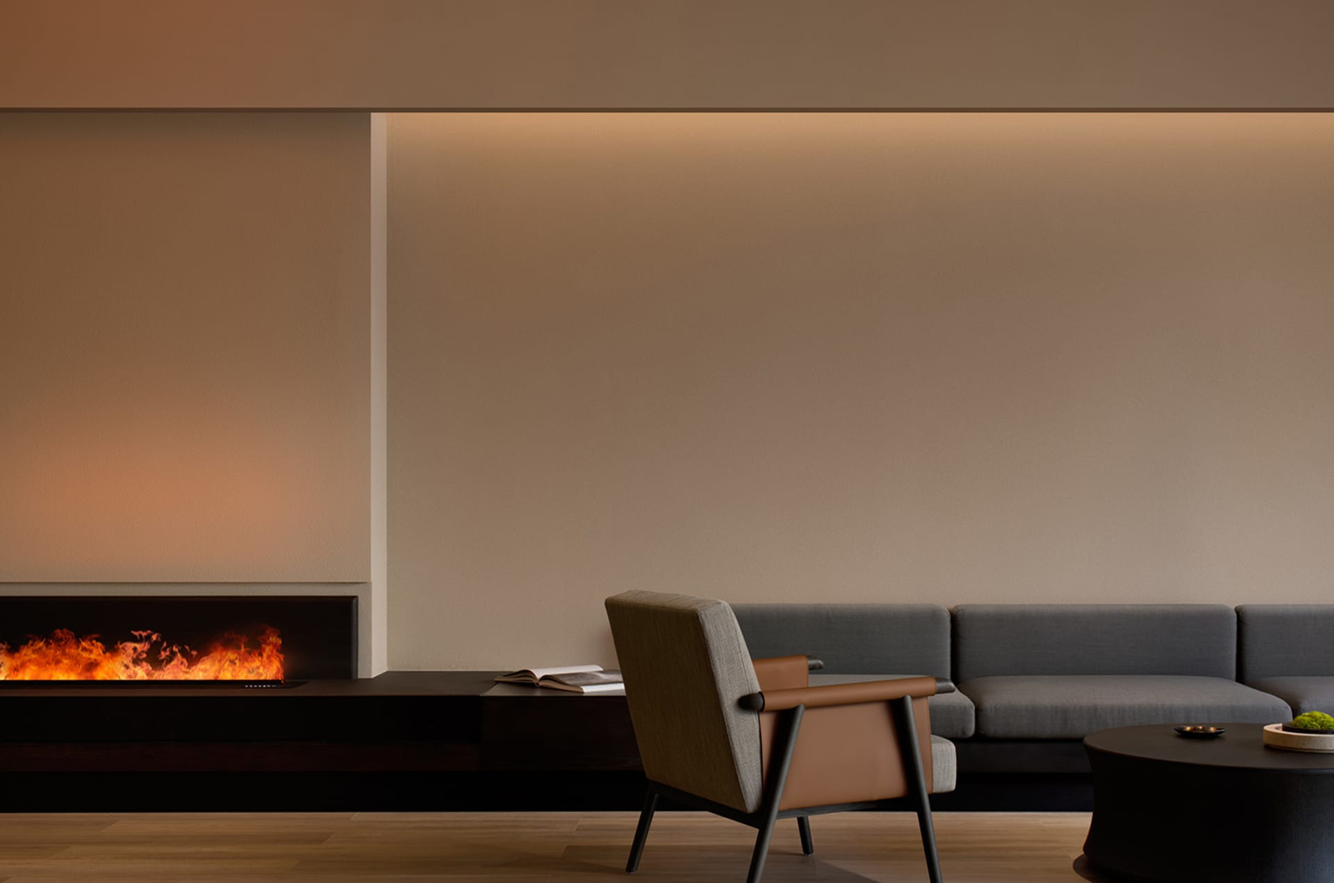 Modern living room with minimalist furniture, a grey sectional sofa, a brown armchair, and a fireplace with a warm ambient li
