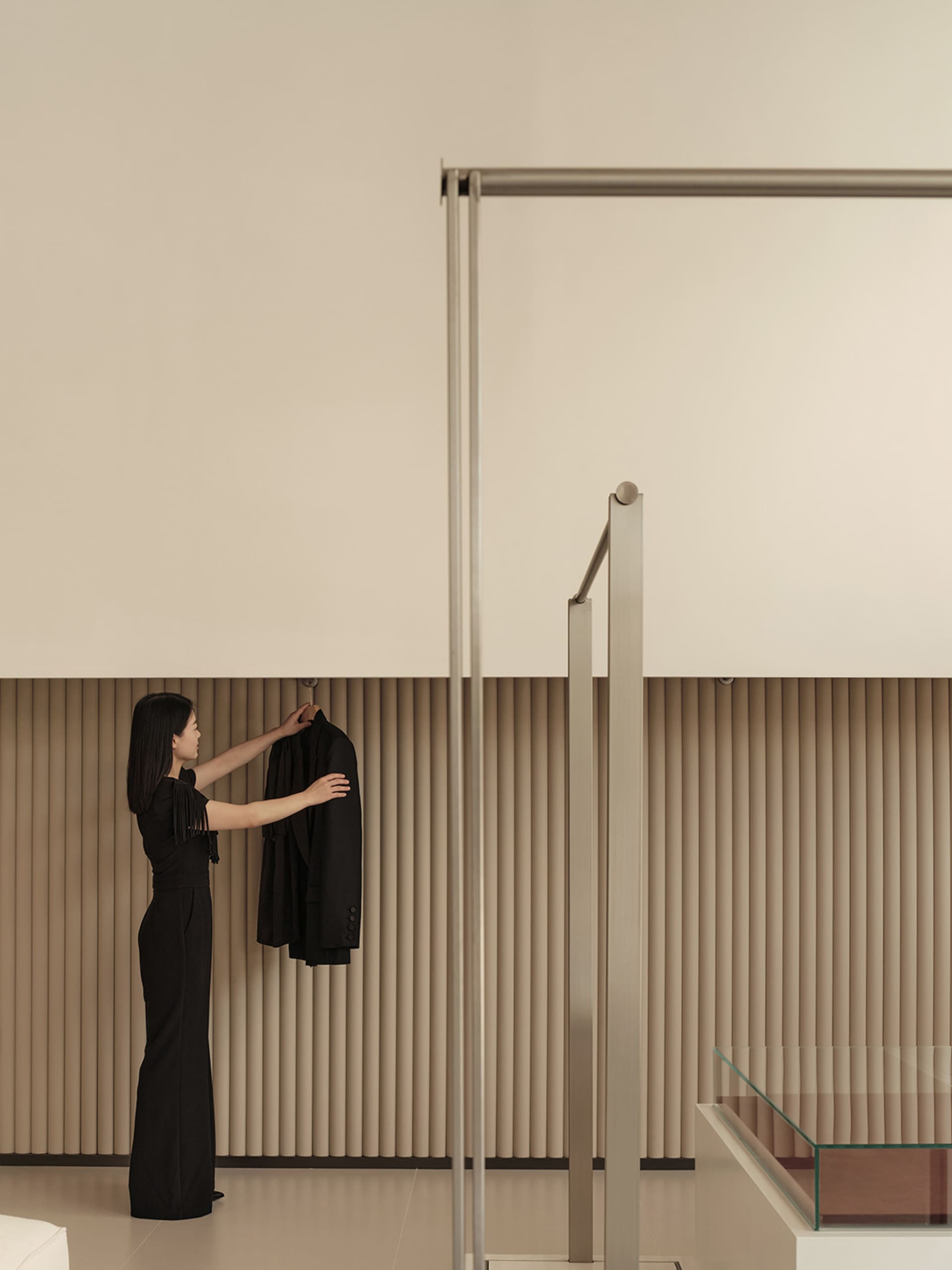 A woman in a minimalist living room, hanging a black coat on a metal drying rack. The room features concrete walls and floor,