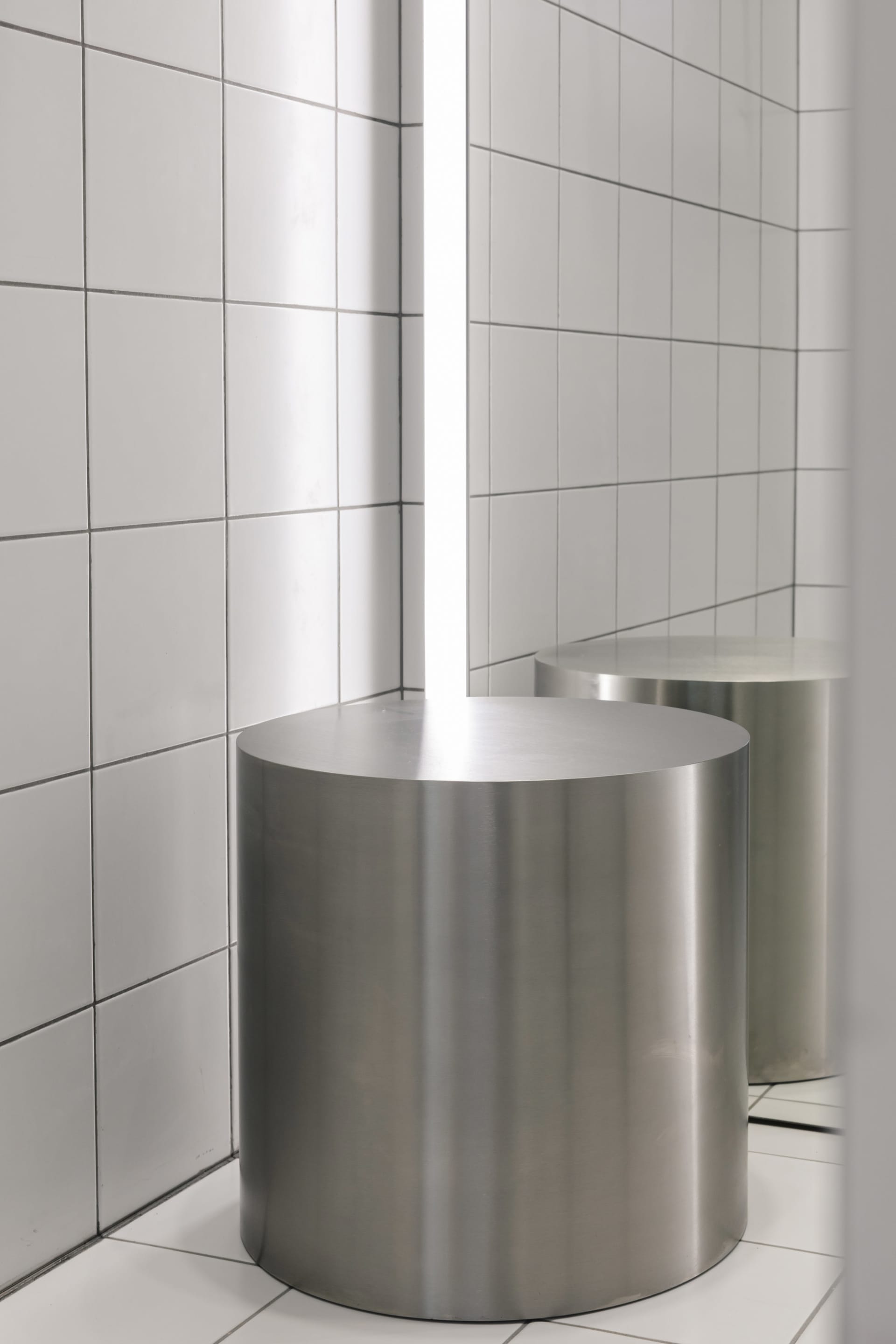Modern minimalist bathroom featuring cylindrical metal stools on a white tiled floor and wall.