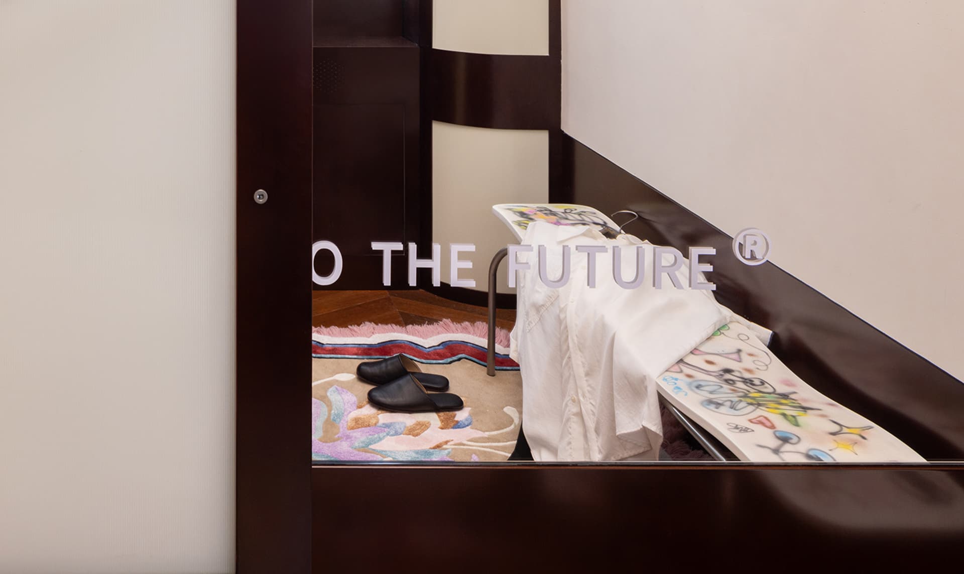 A retail shop display featuring a white shirt and black shoes on a wooden floor with a multicolored rug and a sign reading 'T