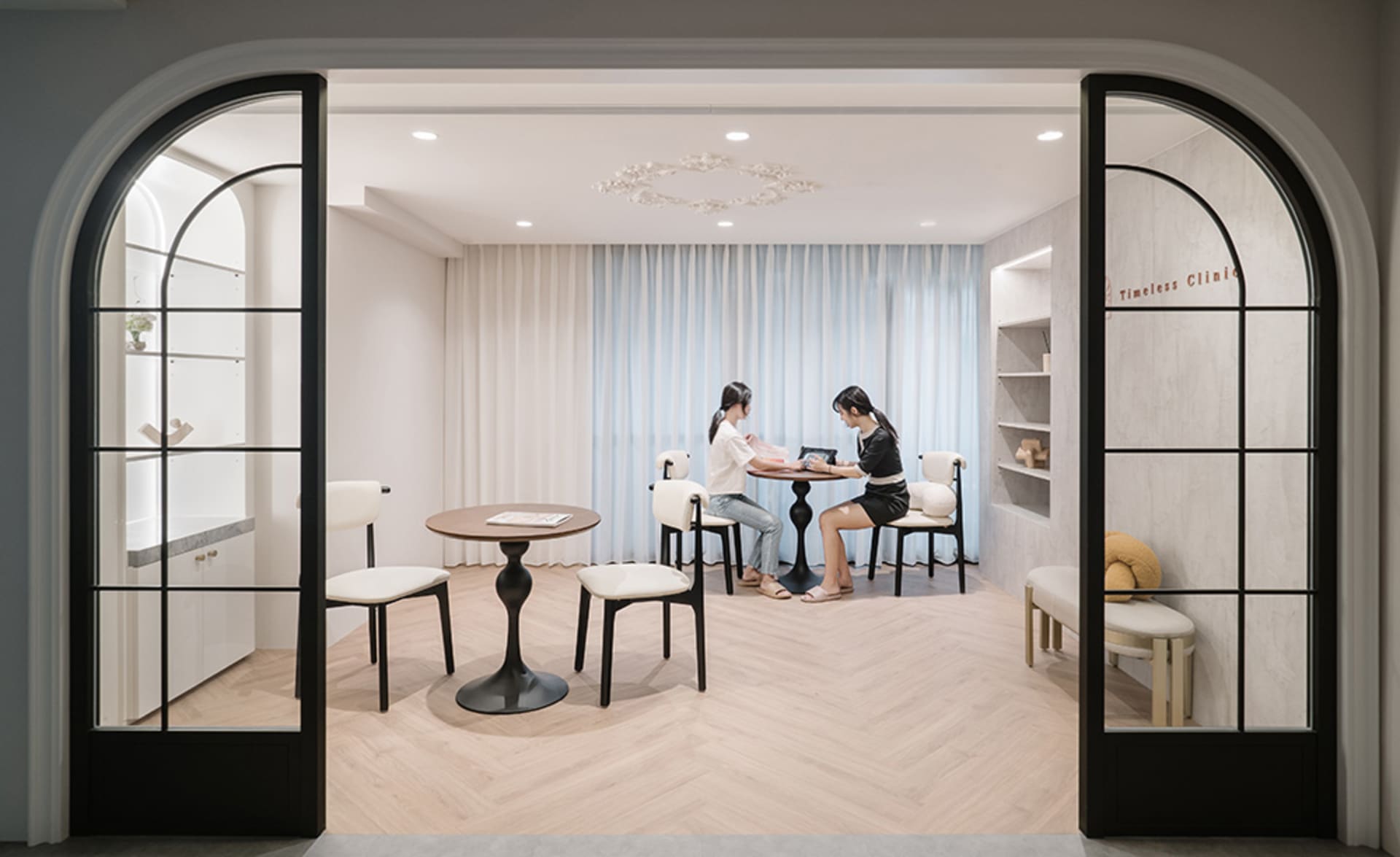 A modern waiting room with light-colored wooden flooring and minimalist decor, featuring two women seated at a table.