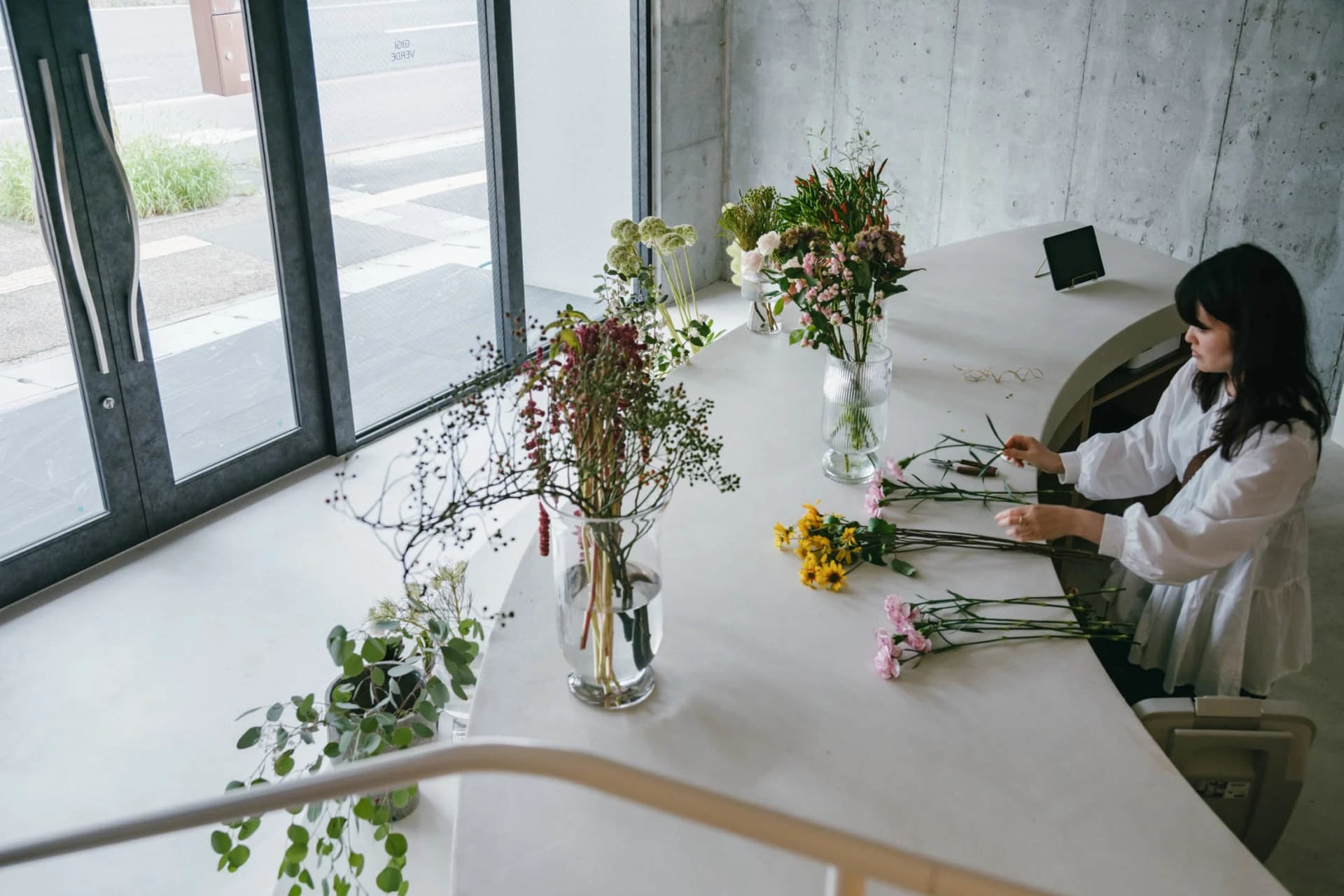A woman sits at a white concrete counter arranging flowers using glass vases.