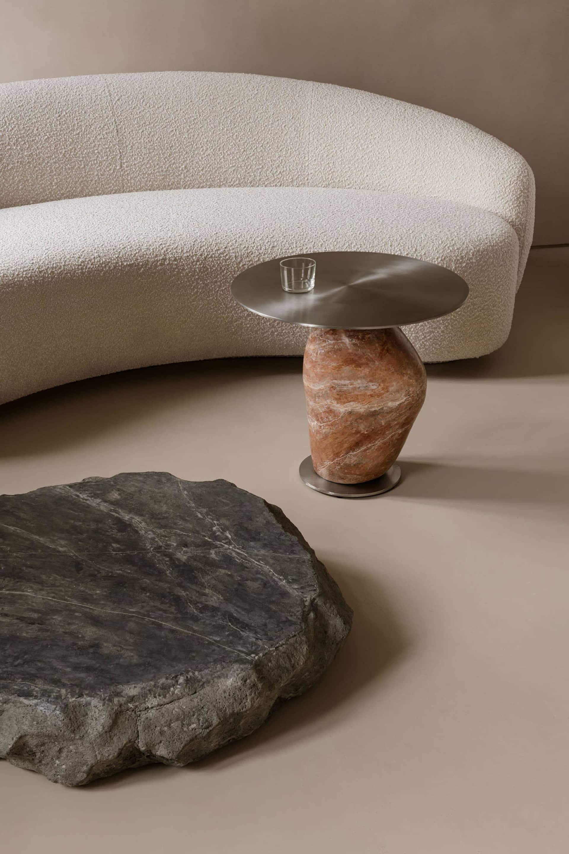 A modern, curved marble side table with a round, metallic top and a vase-like base.