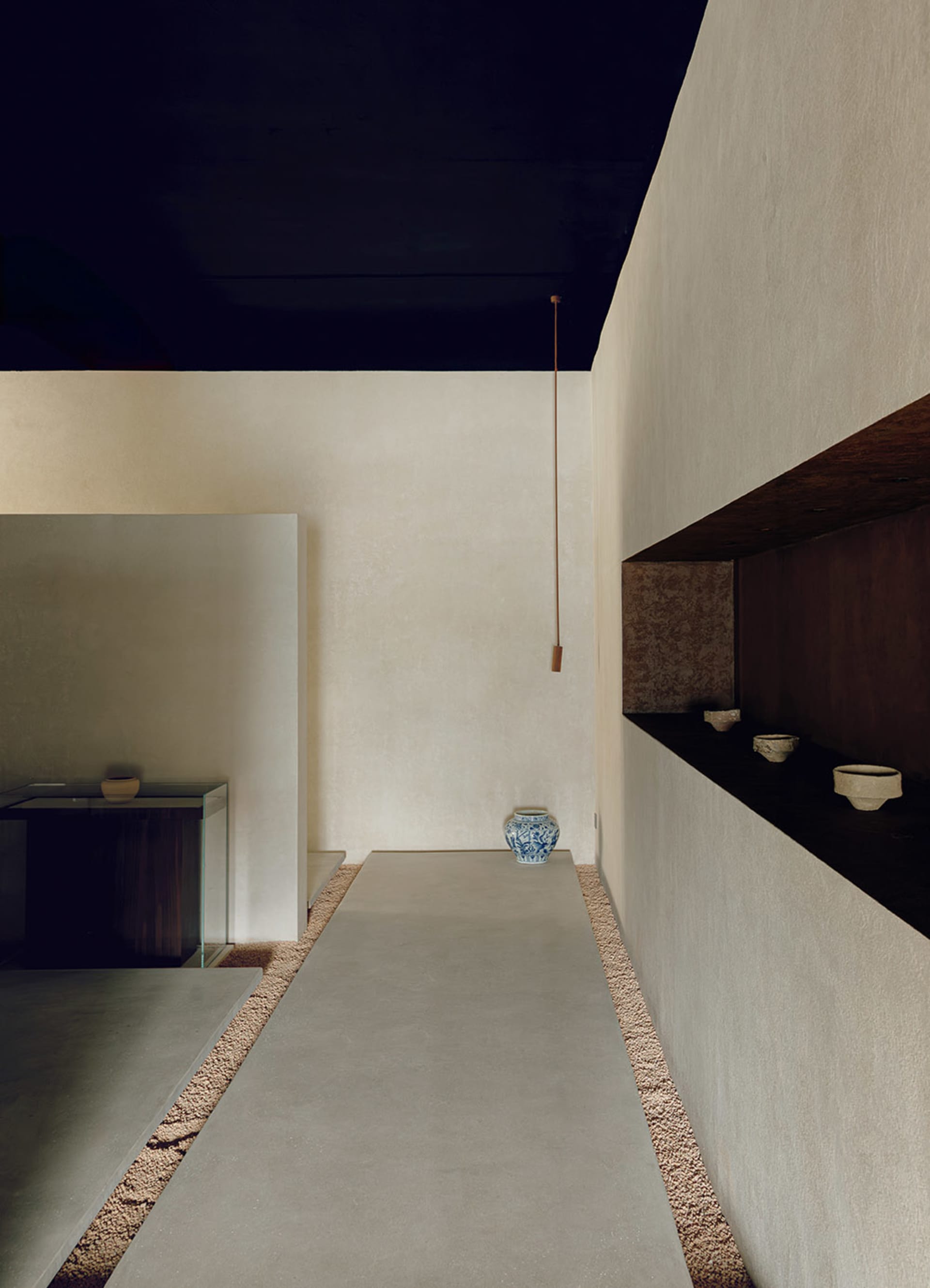 A minimalist living room with concrete walls and floor, featuring a modern shelf with bowls and a blue-and-white vase.