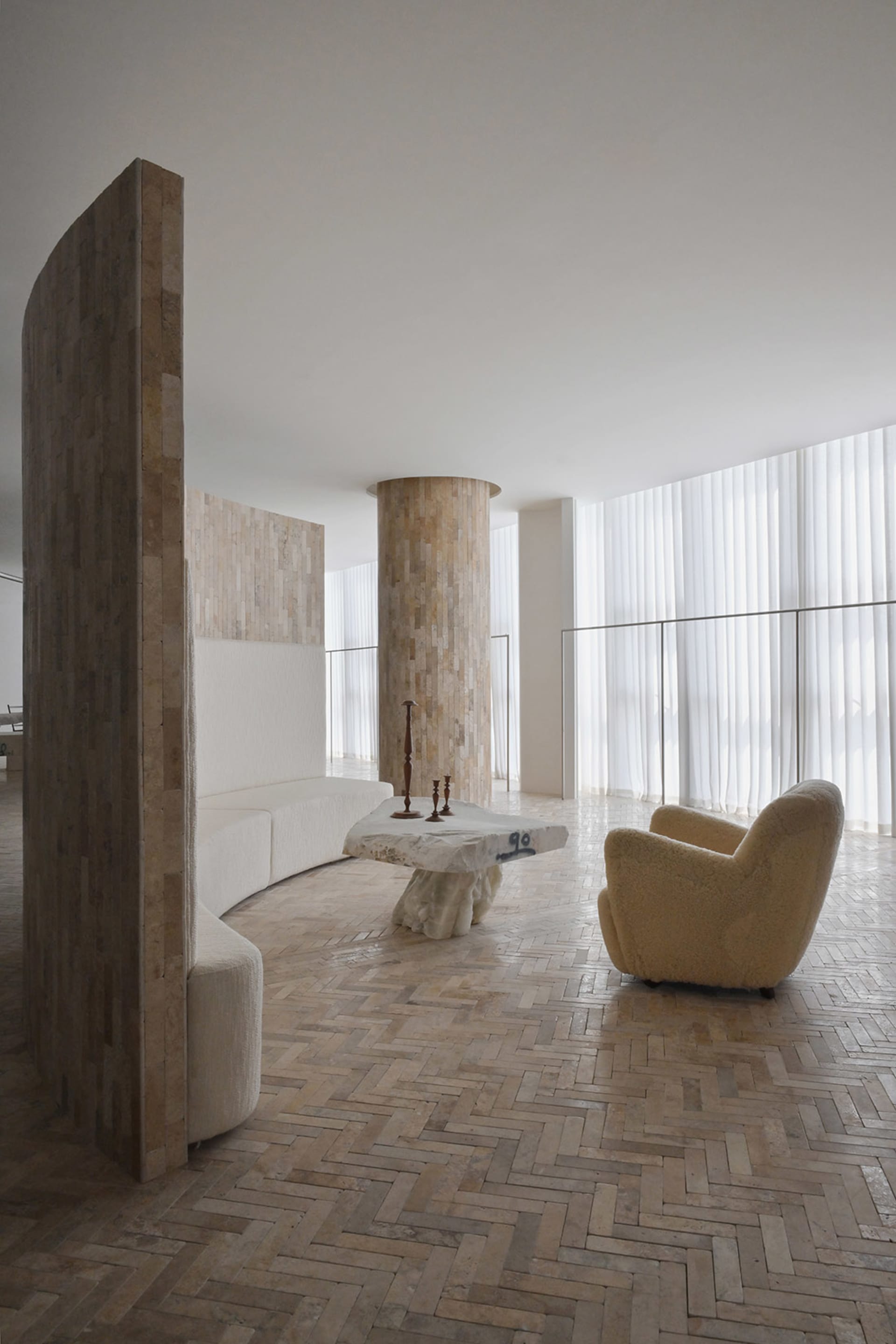 A modern living room featuring a large sofa, a stone table, and a fluffy armchair, set against a light-colored brick wall and