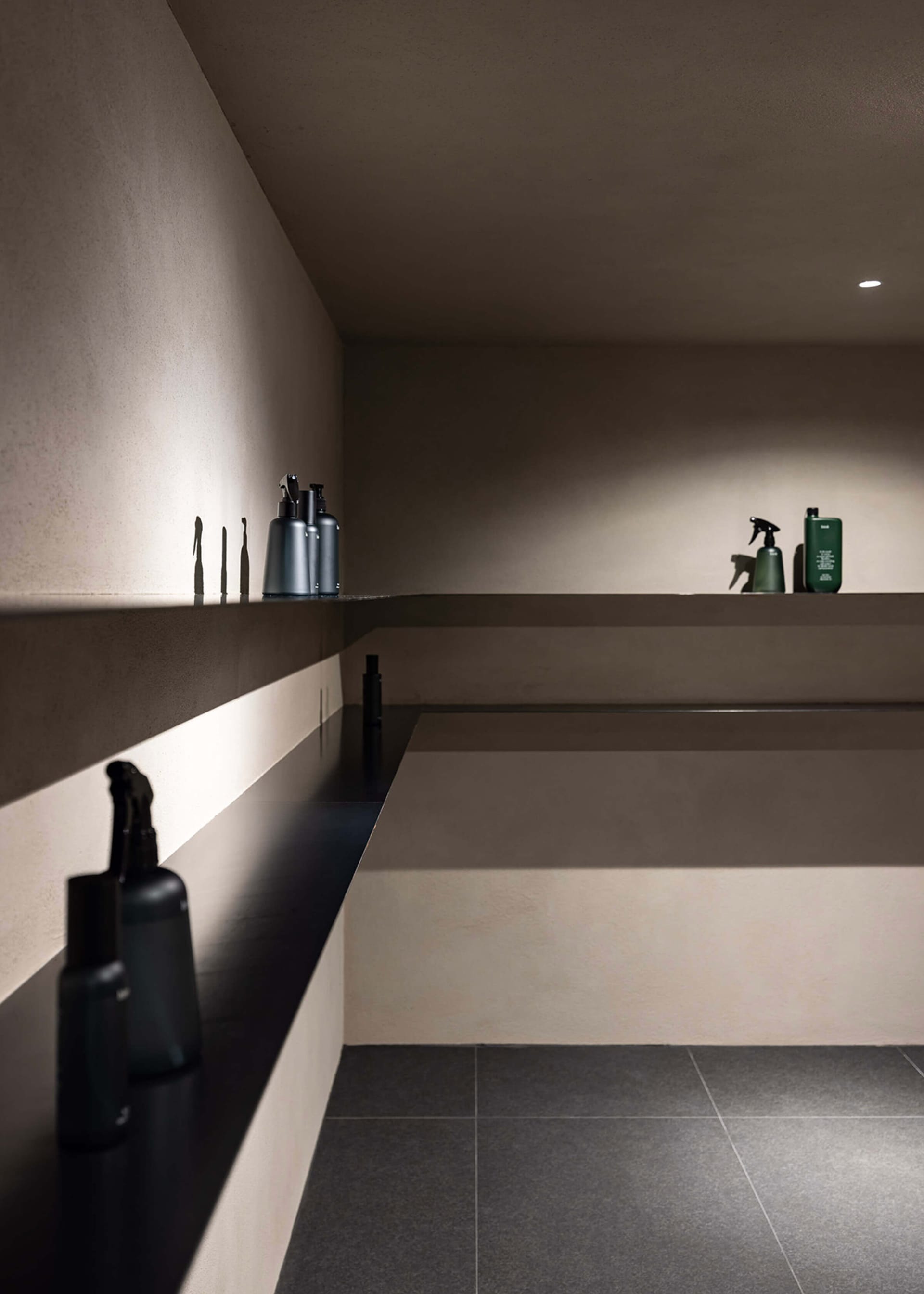 A minimalist bathroom with concrete walls and a black shelf holding various bottles and containers.