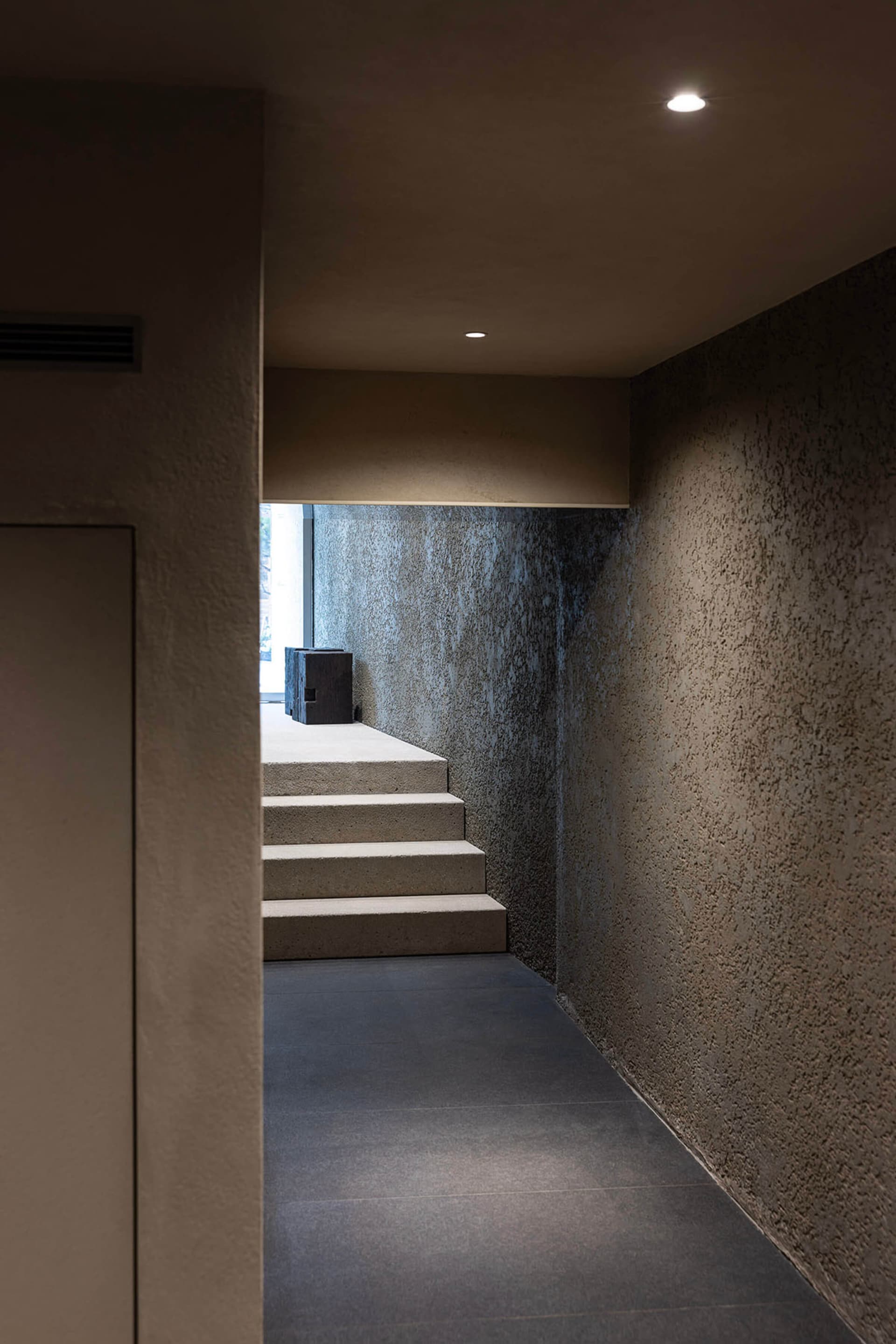 A modern, minimalist hallway with concrete stairs and walls, illuminated by recessed lighting.