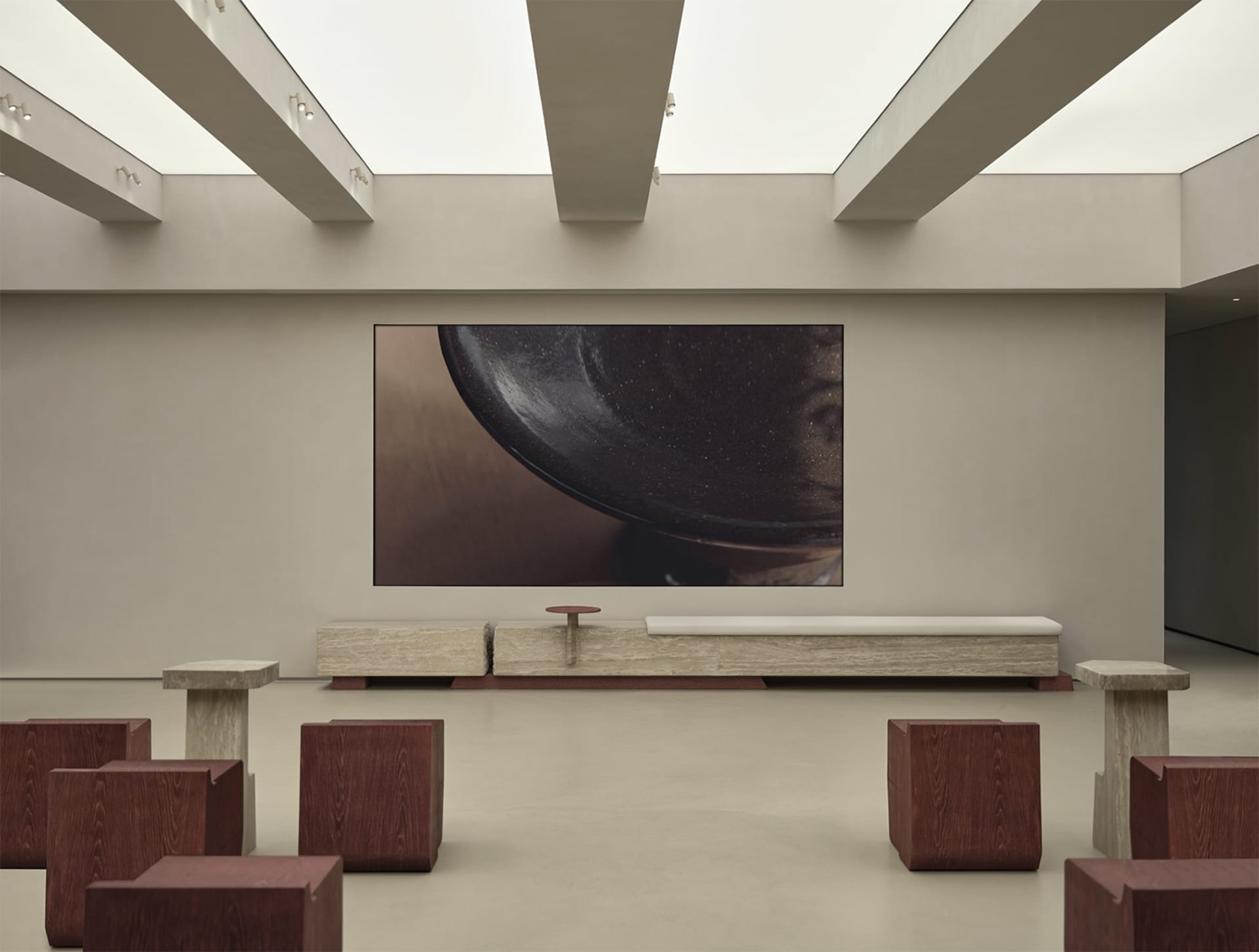 A minimalist concrete lobby with seating, a low bench, and a large screen displaying a bowl.