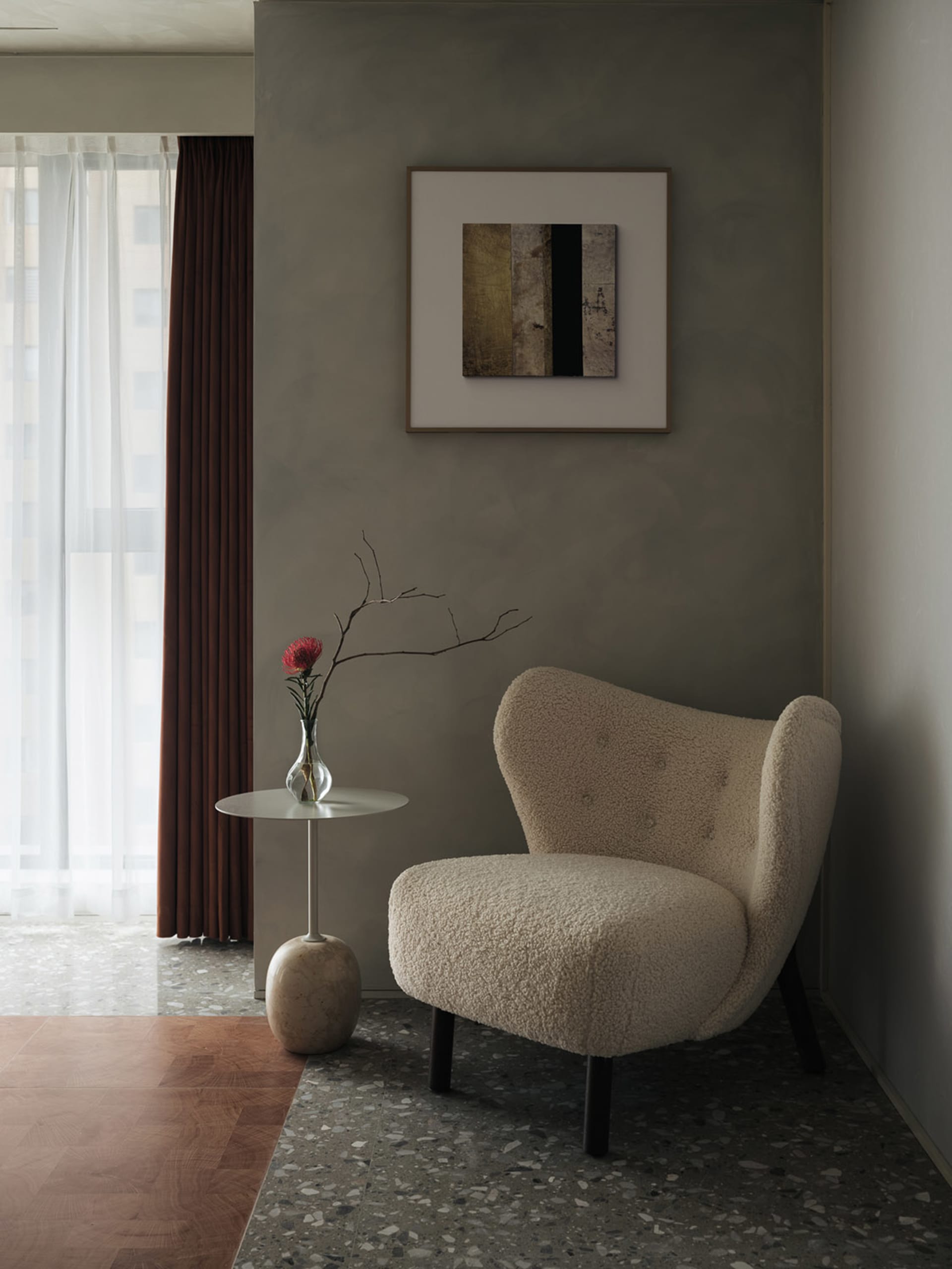 A minimalist living room featuring a beige cushioned armchair, a small marble side table with a red carnation, and a framed a