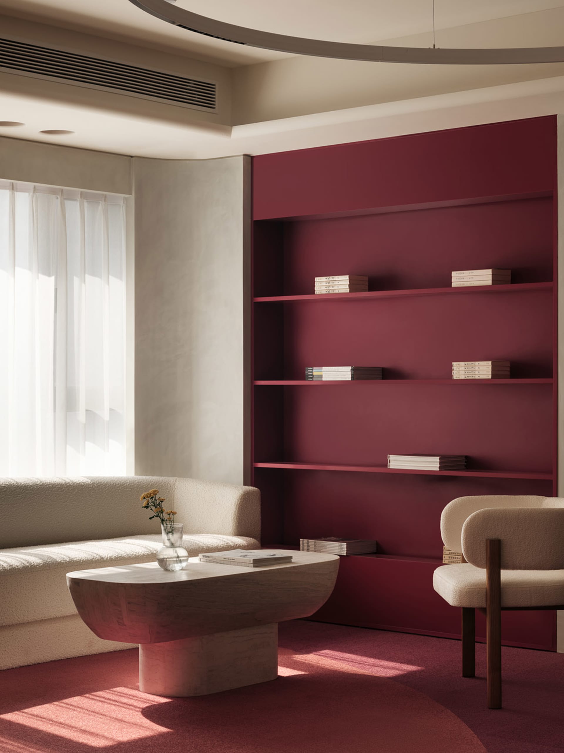 A minimalist living room with a red shelving unit and white furniture, featuring a table, a chair, and a window with sheer cu