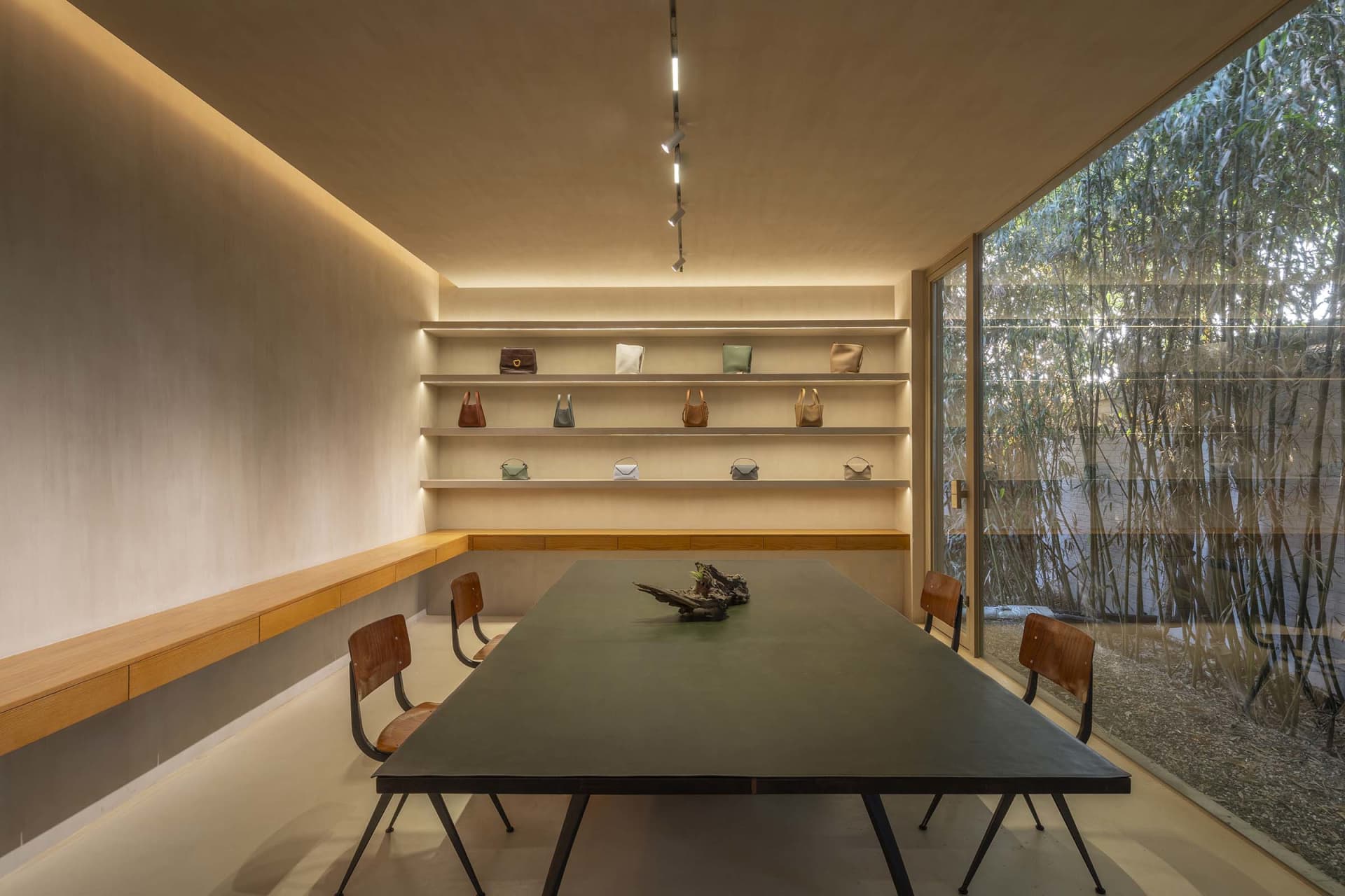 A modern conference room featuring a table, chairs, and shelves with bags, surrounded by a bamboo garden.
