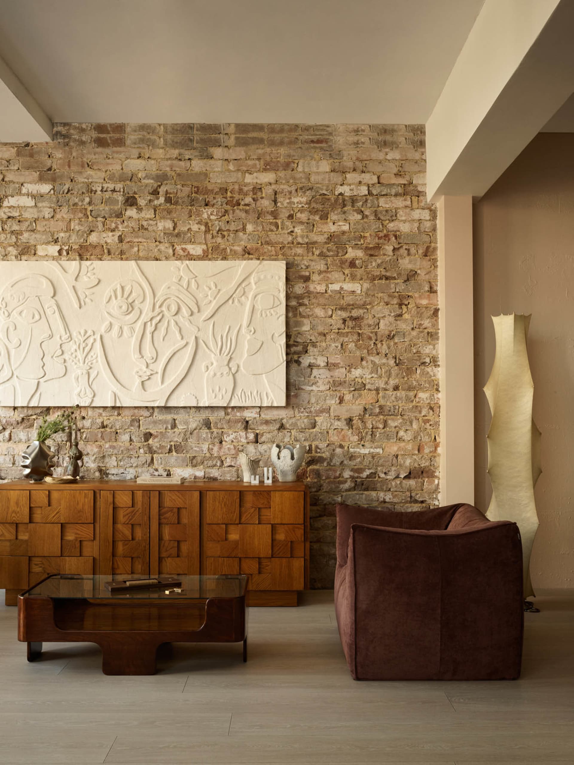 Modern living room featuring a brick wall, wooden cabinet, and a brown armchair with a geometric floor lamp.