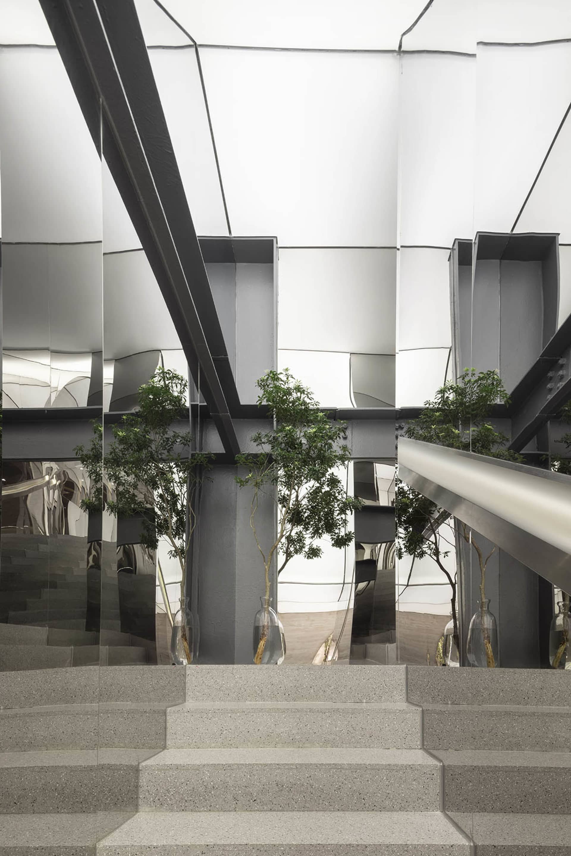 A modern interior design featuring concrete stairs with a metal handrail, surrounded by glass panels with green plants.